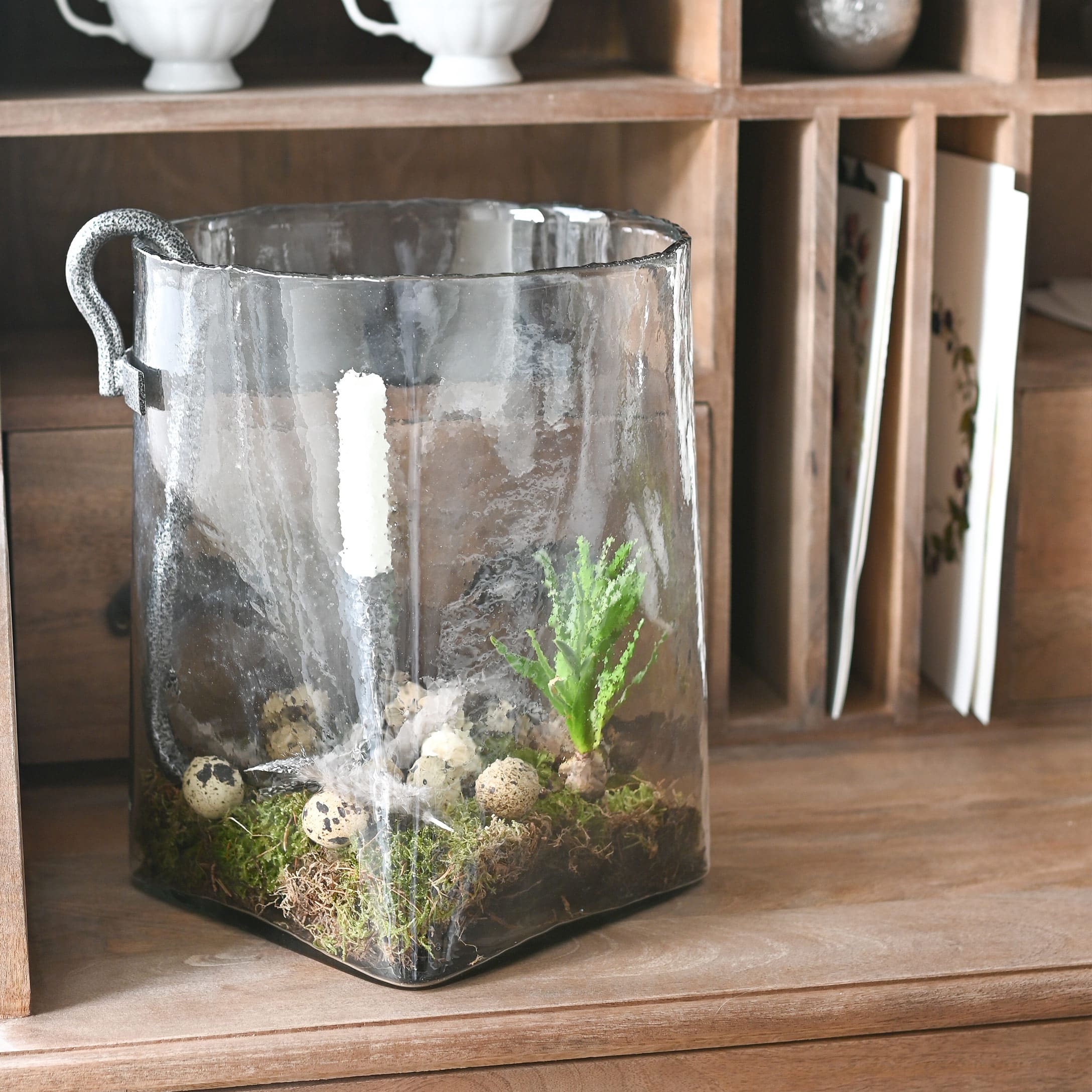 Transparenter Glasbehälter mit Griff, dekoriert mit Moos und Wachteleiern auf einem Holztisch.
