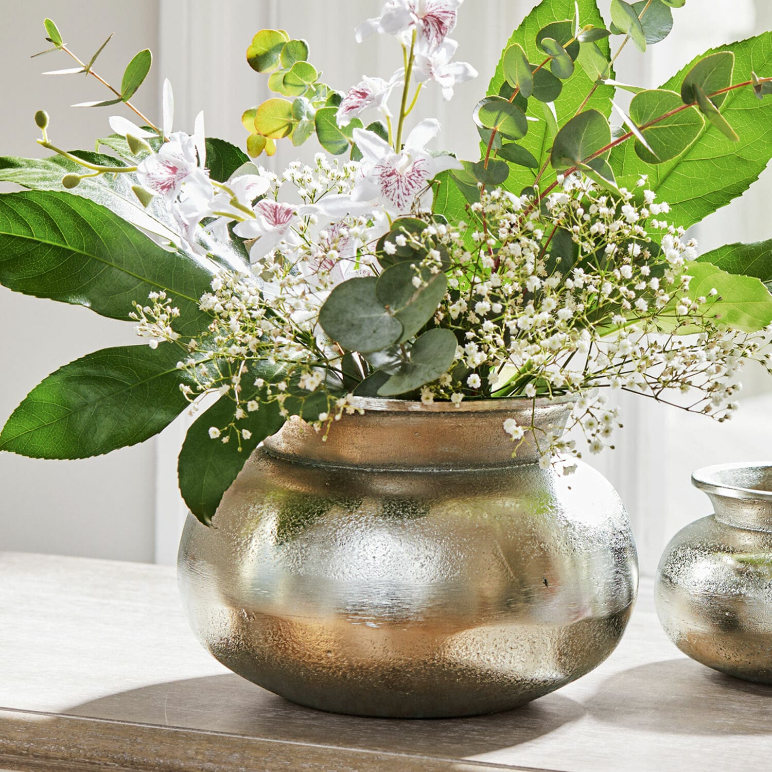 Silberne Metallvase mit weissen Blumen und grünen Blättern auf Holztisch, Wohnaccessoire
