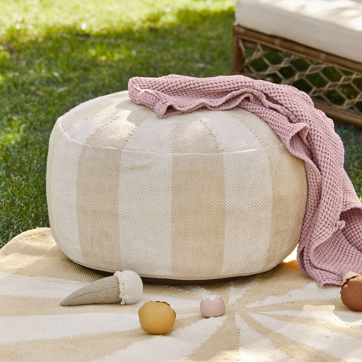 In- & Outdoor-Pouf Wobbilo Beiger runder Stoff-Pouf mit rosa Strickdecke und Spielzeug auf Gras im Garten