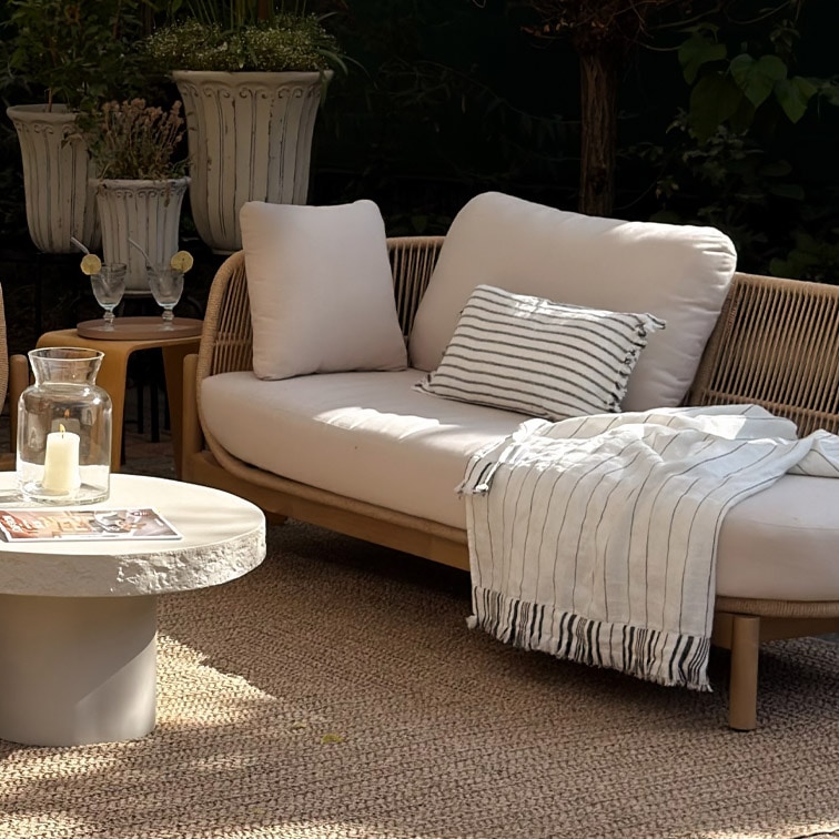 Moderne Gartensofa mit hellen Kissen und gestreiftem Plaid auf natürlichem Teppich im Garten
