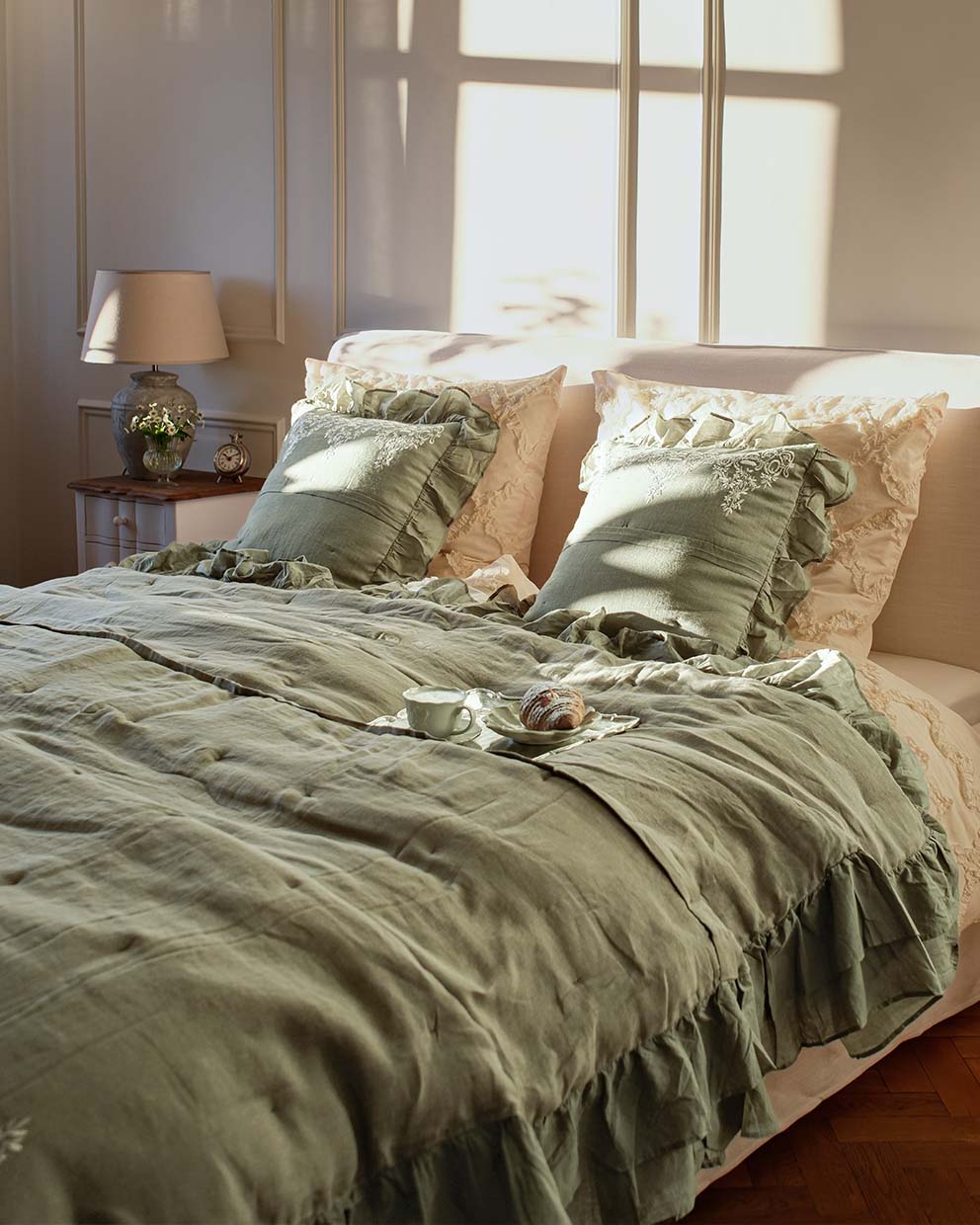 Grüne Leinenbettwäsche mit Rüschen und bestickten Kissen in hellem Schlafzimmer mit Lampe und Uhr