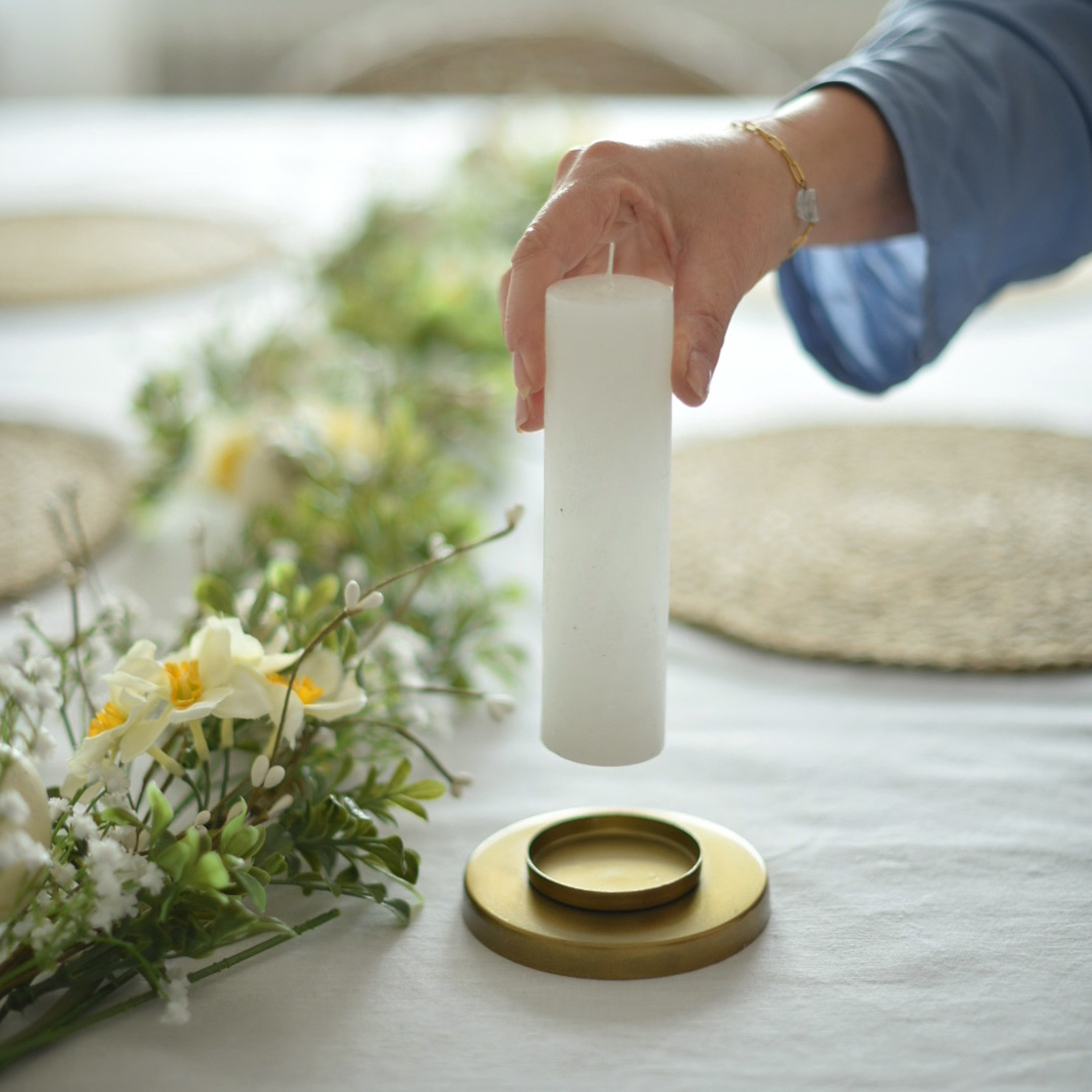 Weiße Stumpenkerze über goldenem Kerzenhalter mit Blumenarrangement auf weissem Tischtuch