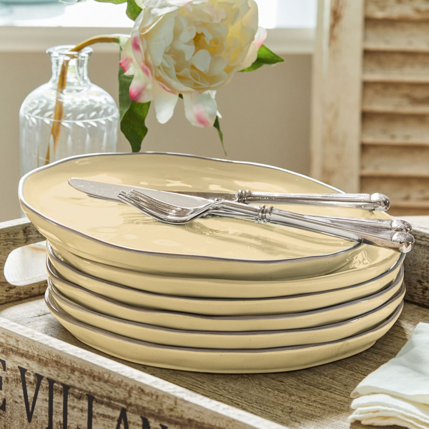 Set aus cremefarbenen Keramiktellern mit metallischem Rand und silbernem Besteck auf Holztablett