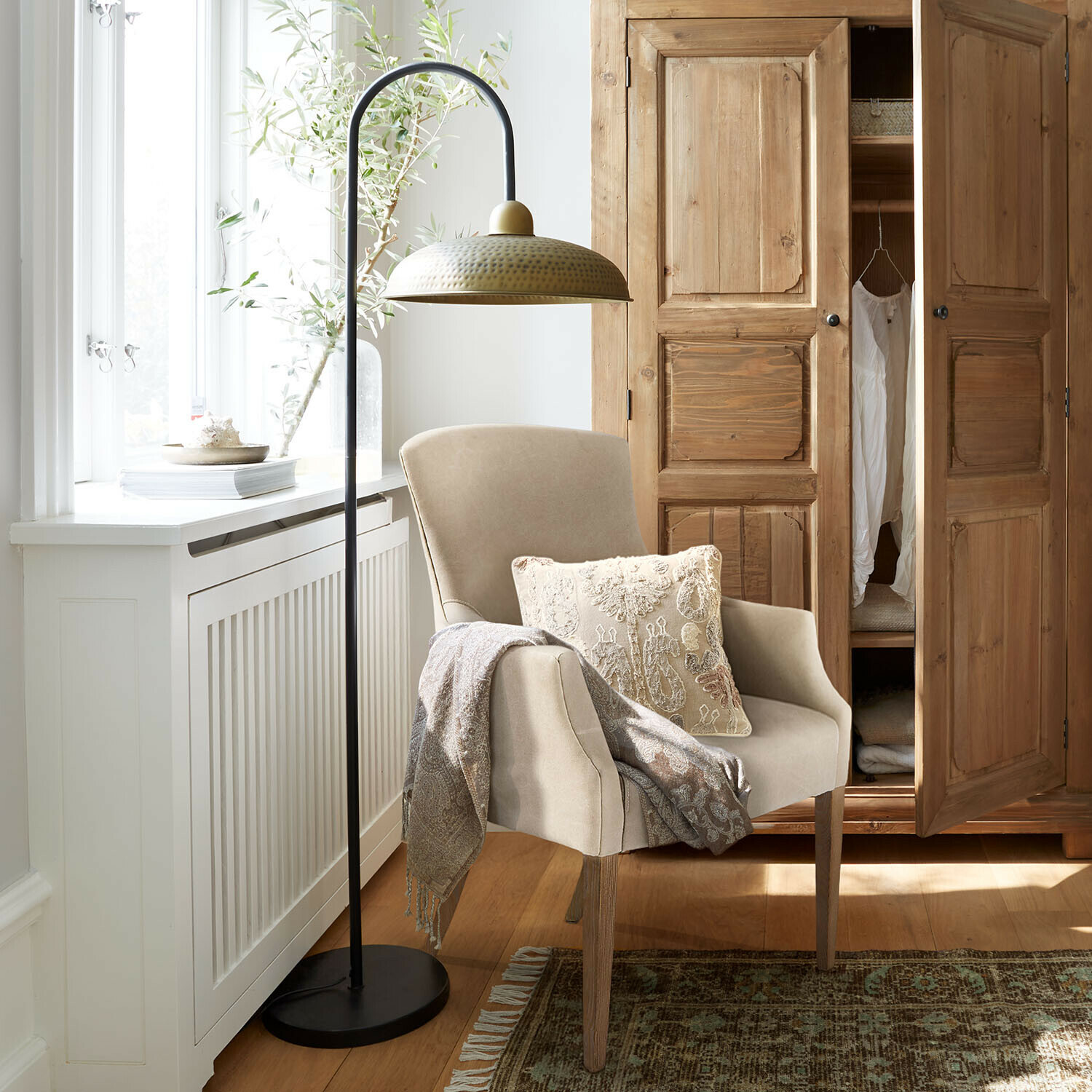 Beiger Sessel mit Holzbeinen, dekorativem Kissen und Decke, daneben schwarze Stehlampe mit messingfarbenem Schirm