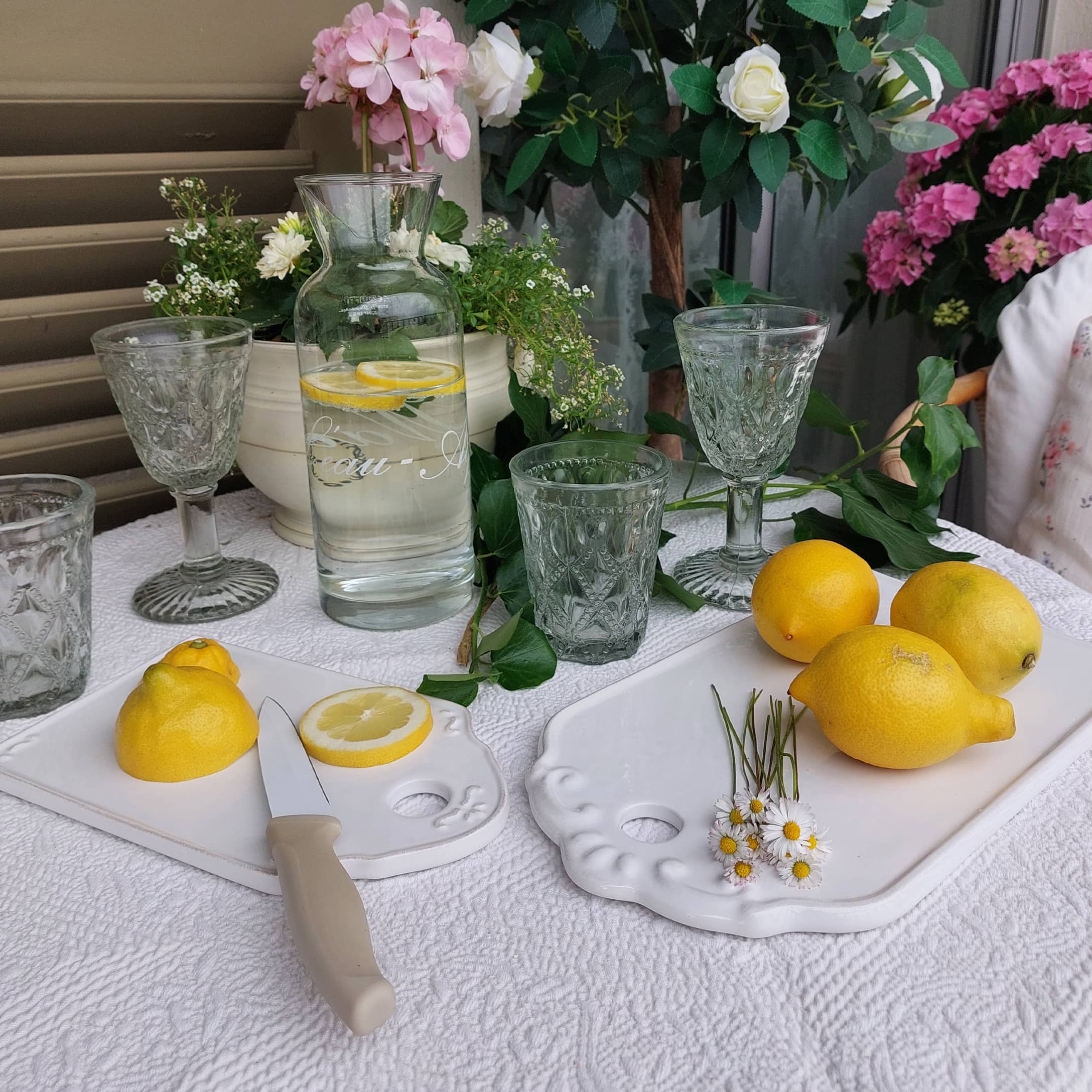 Glaskaraffe mit Zitronenwasser auf weisser Tischdecke, dekorative Gläser und weisse Keramik-Schneidebretter