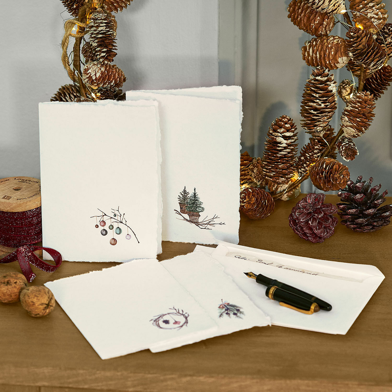 Grusskarte 4er Set Velorine Set Weihnachtskarten aus Papier mit Tannen- und Zapfenmotiven auf Holztisch mit Füllfederhalter und Dekoration