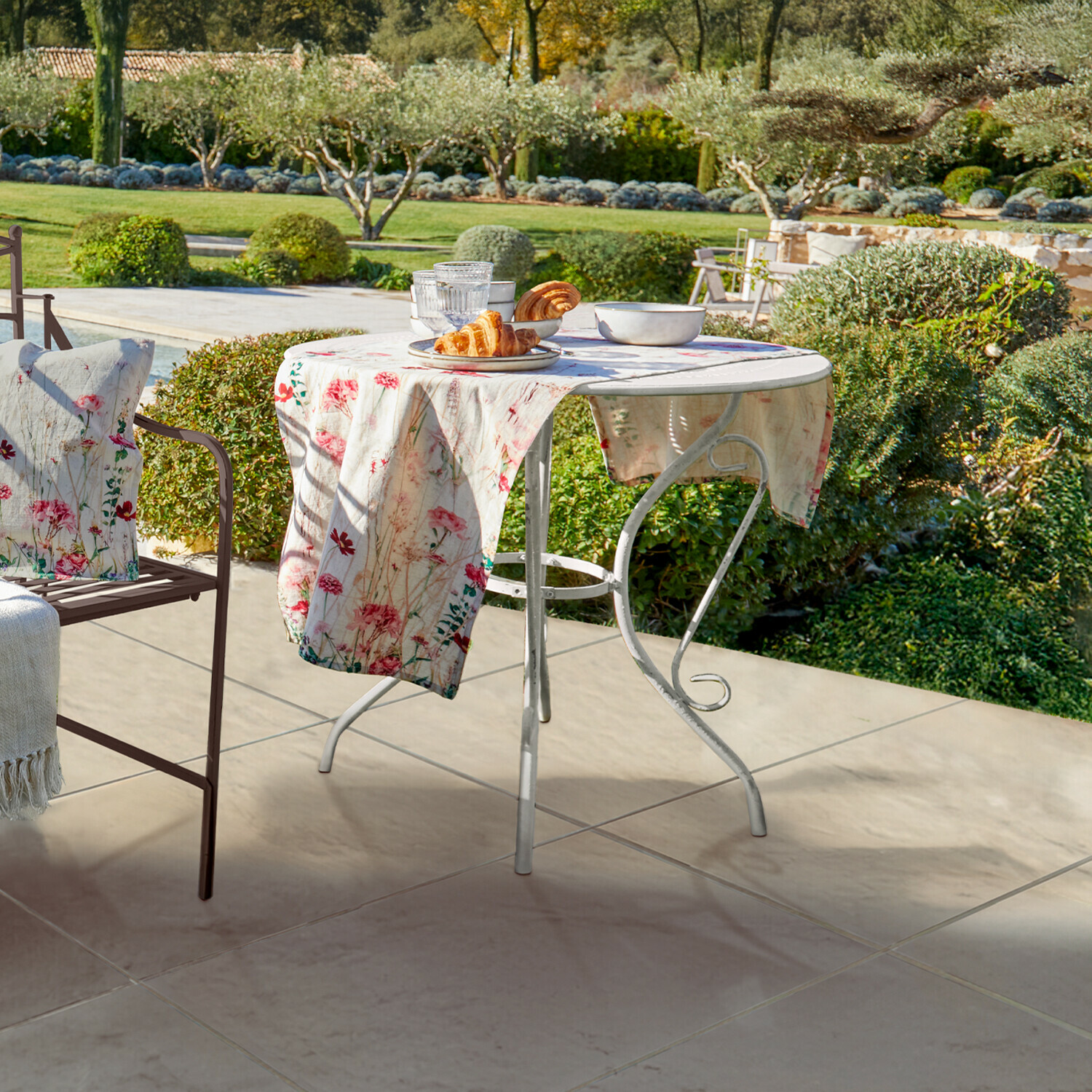 Tisch Aube Runder weisser Metall-Gartentisch mit floralem Tischtuch und Gebäck auf Terrasse mit Gartenblick