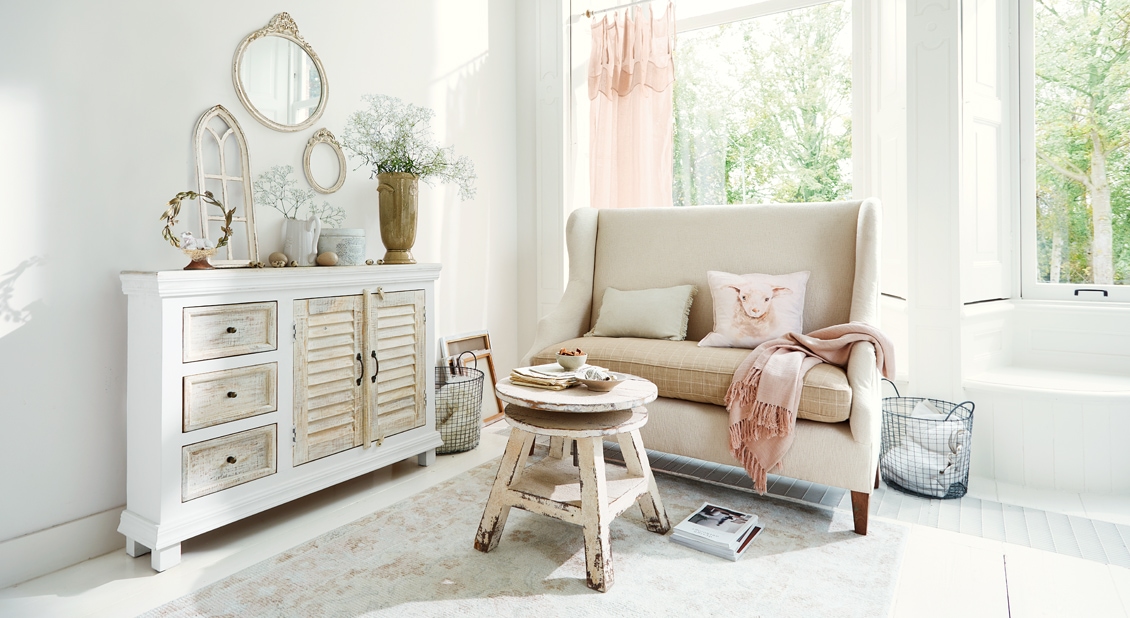 Gemütliches Wohnzimmer im Shabby-Chic mit weissem Holzsideboard und beigem Sofa mit Kissen und rosa Decke