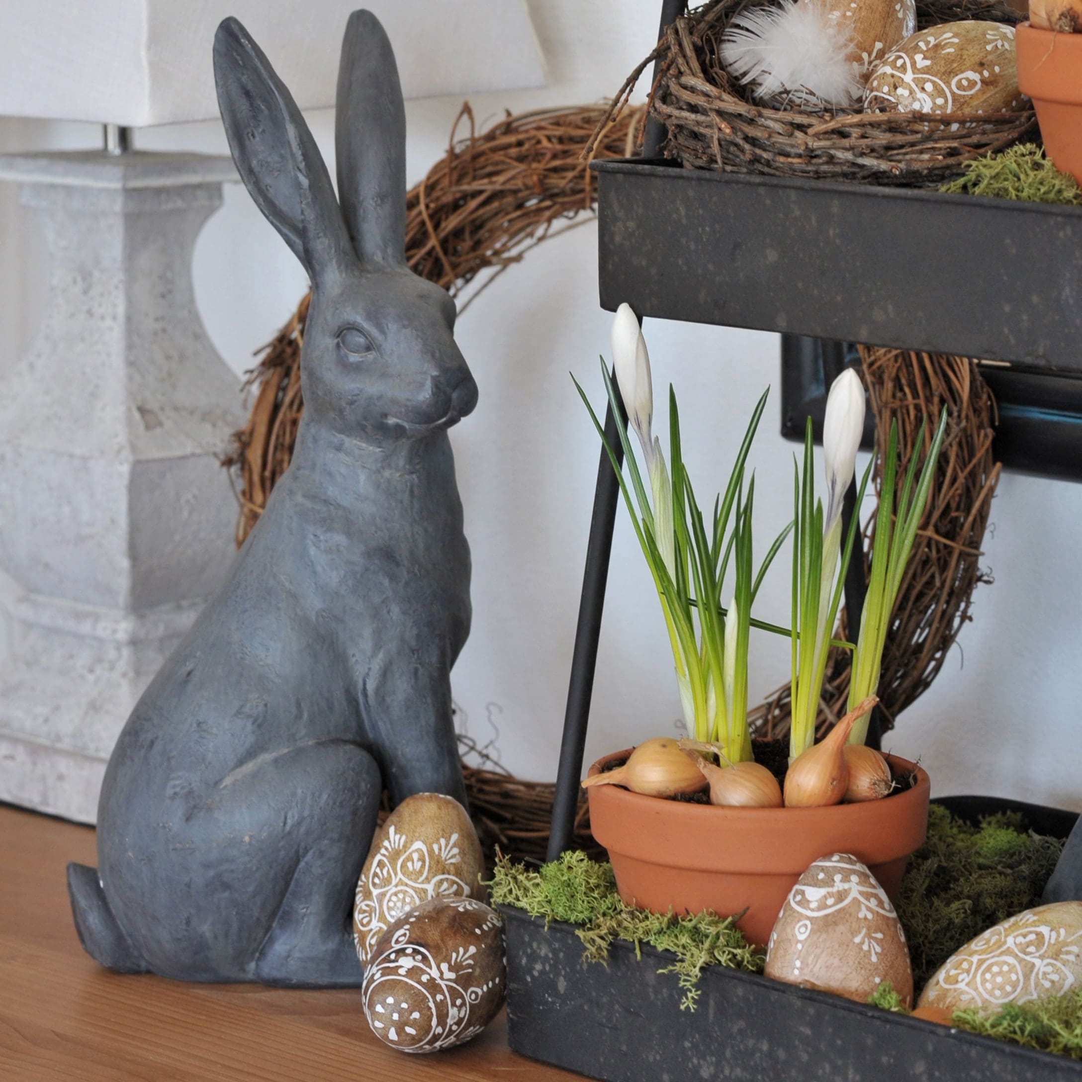 Graue Keramik-Hasenfigur neben Blumentopf mit weissen Blüten und dekorativen Ostereiern für Frühlingsdekoration