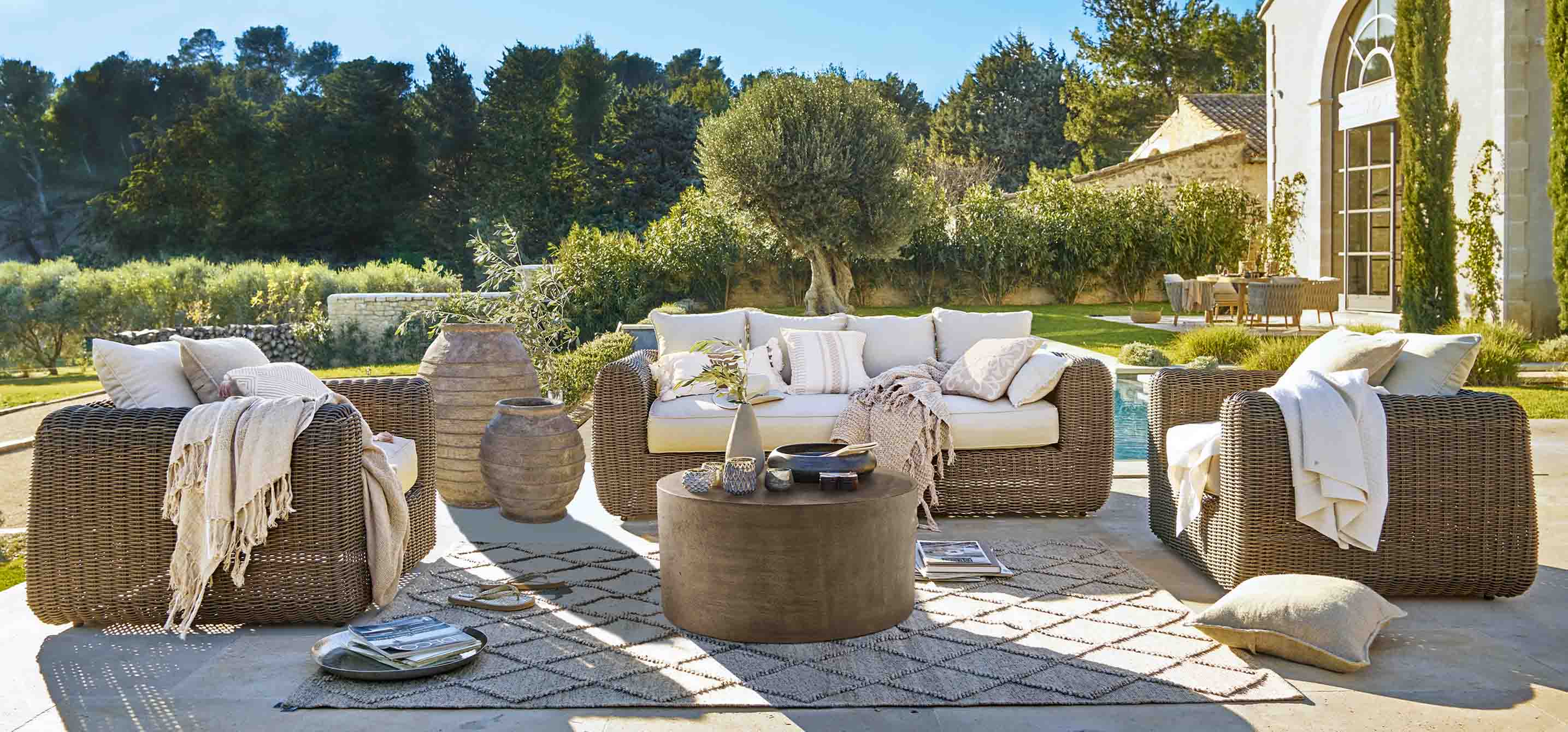 Braunes Rattan-Gartenmöbel mit cremefarbenen Kissen und rundem Tisch auf sonniger Terrasse