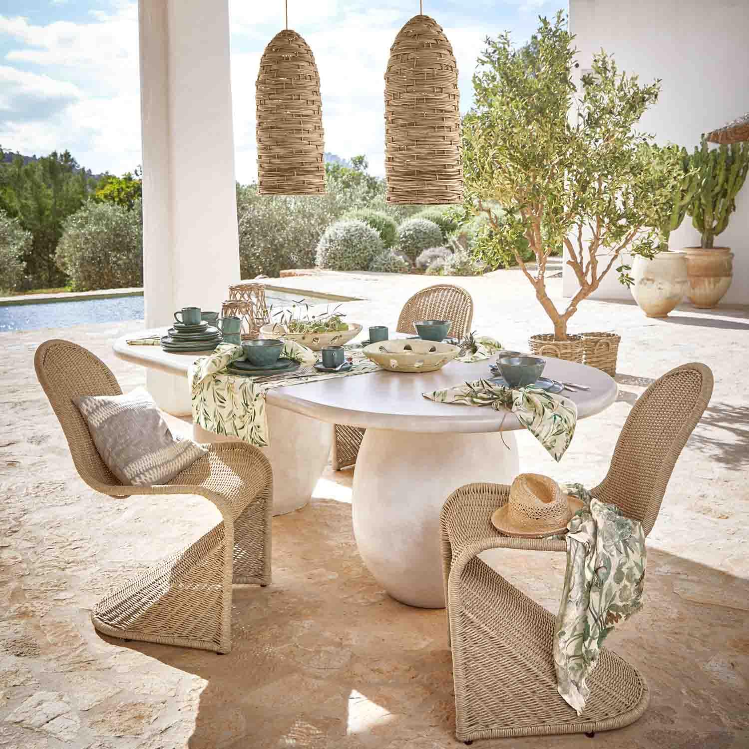 Ovaler weisser Steintisch mit Rattanstühlen und natürlichen Hängeleuchten in sonnigem Garten