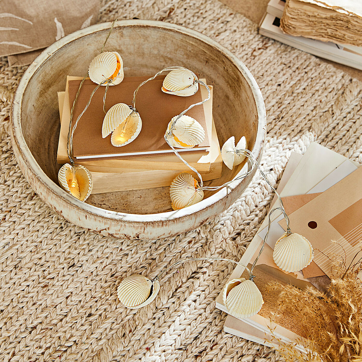 Lichterkette mit Muscheln in Naturtönen auf beigem Teppich mit Holzschale und Büchern dekoriert