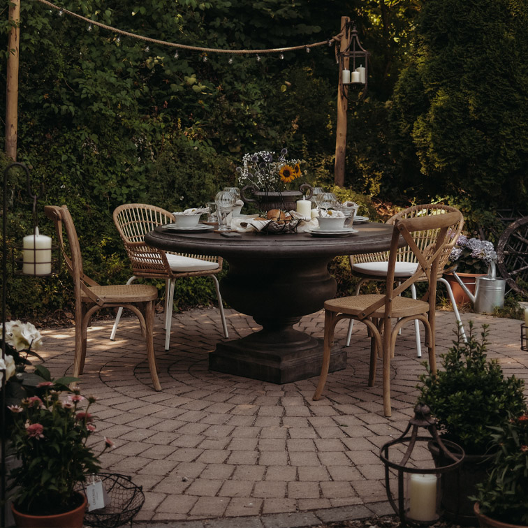 Runder Gartentisch aus dunklem Holz mit Rattanstühlen auf gepflasterter Terrasse mit Kerzen und Blumen dekoriert