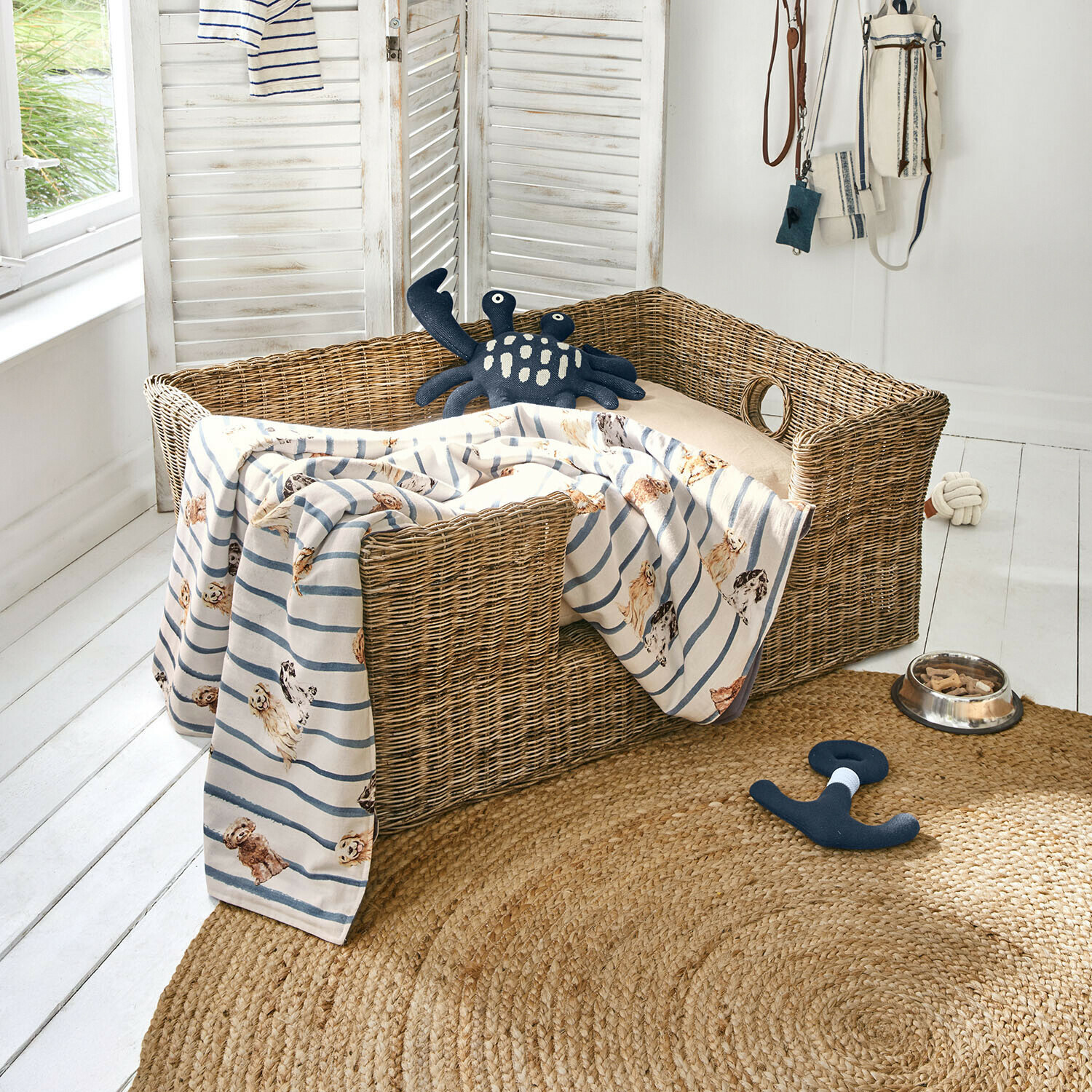 Haustierkorb Knuddo Weiden-Hundebett mit beigem Kissen und gestreifter Decke mit Hunde-Motiv, Spielzeug und Futternapf auf dem Boden