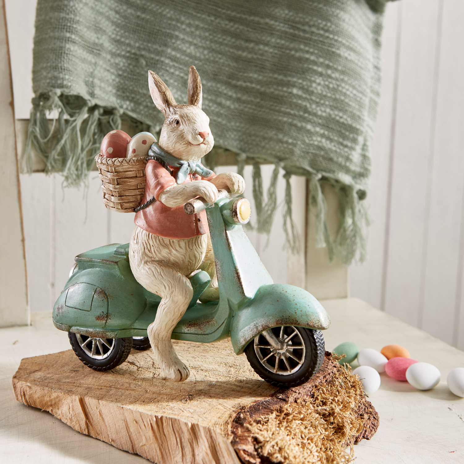 Hasenfigur auf grünem Roller mit Korb voller bunter Ostereier auf Holzsockel