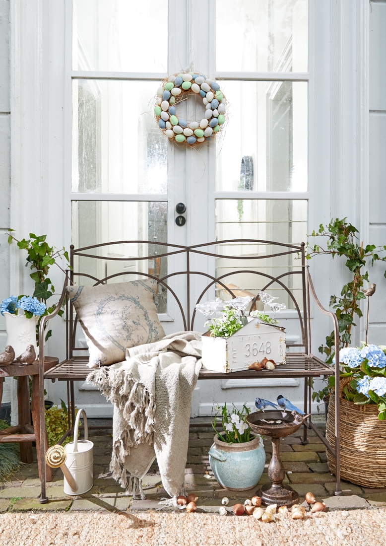 Metall-Gartenbank mit beigem Plaid und Kissen, Blumenarrangements und pastellfarbener Eiergirlande