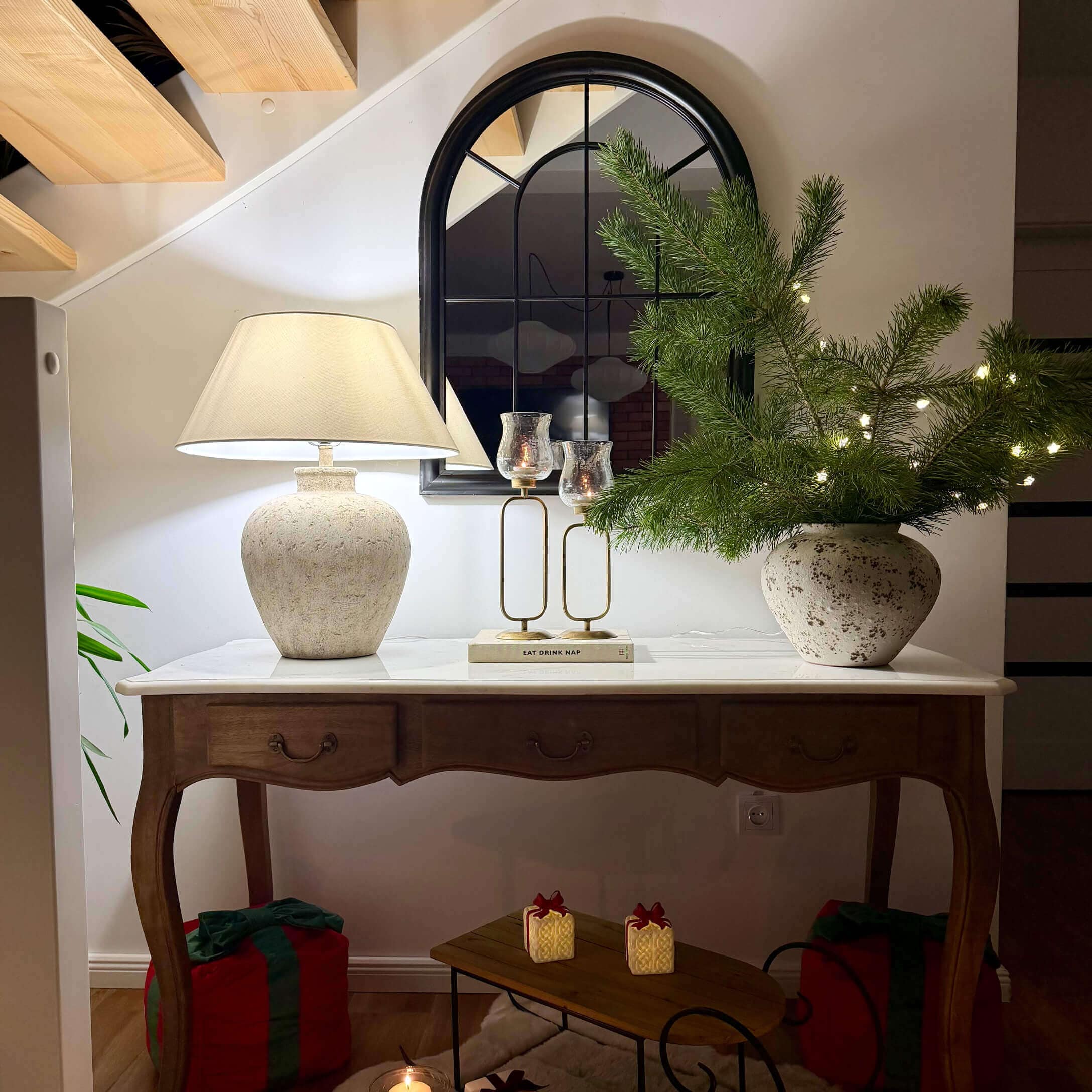Holztisch mit weissem Tischblatt, Keramiklampe, Spiegel und Weihnachtsdekoration mit grünen Zweigen und Lichterkette