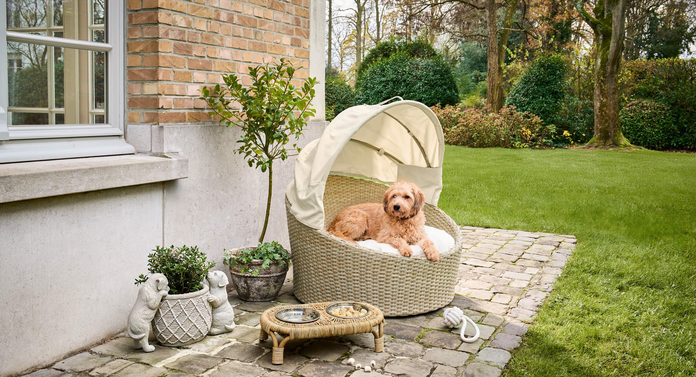 Beiger geflochtener Hundekorb mit Dach auf Stein-Terrasse, gemütlicher Platz im Garten