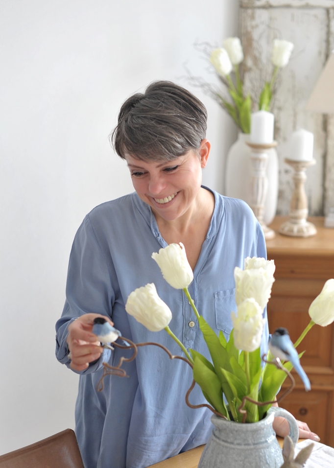 Frau im blauen Hemd mit weissen Tulpen und dekorativen Vögeln in einem grauen Krug auf dem Tisch