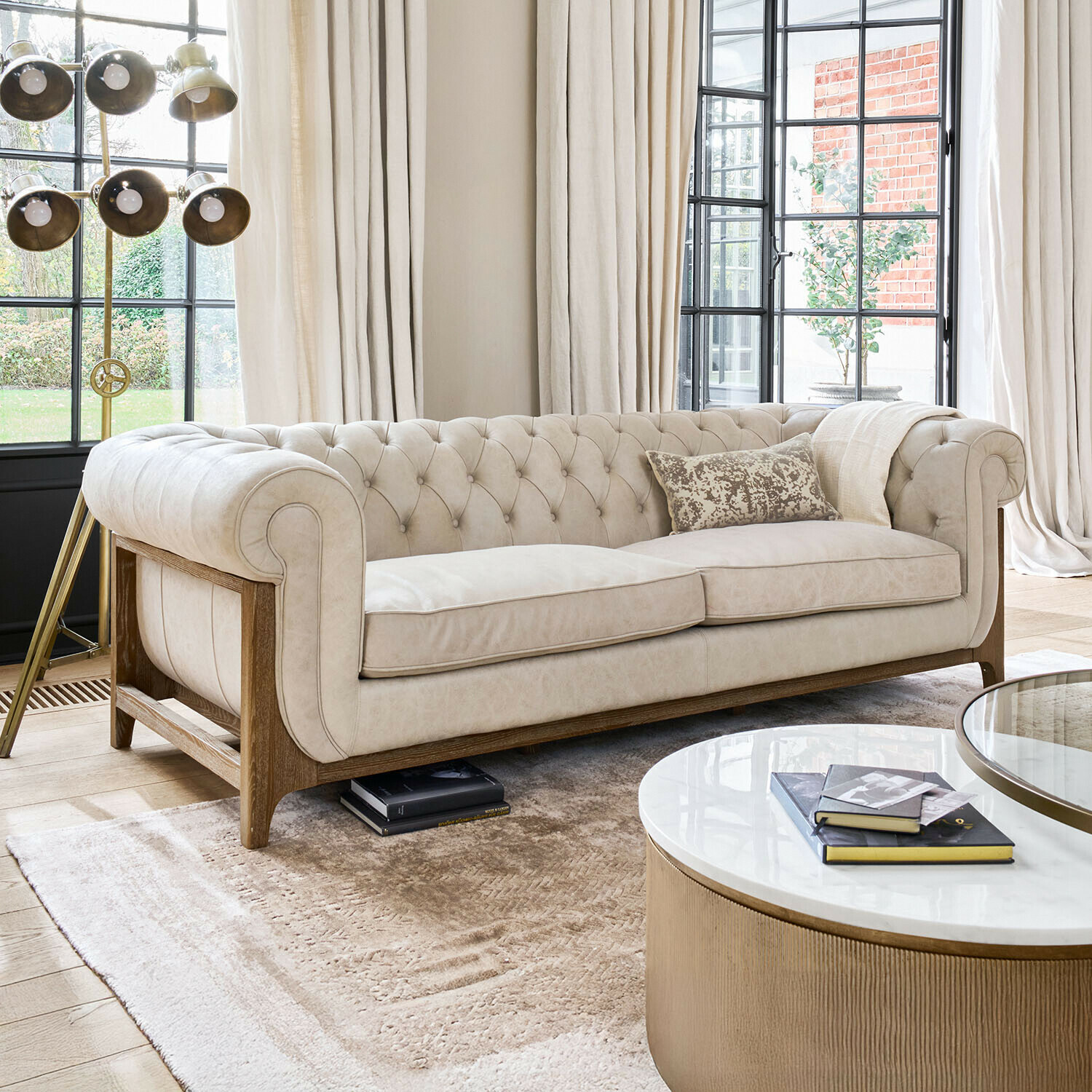 Sofa Swansboro Beiges Chesterfield-Sofa mit Holzrahmen in hellem Wohnzimmer mit grossen Fenstern und Stehleuchte
