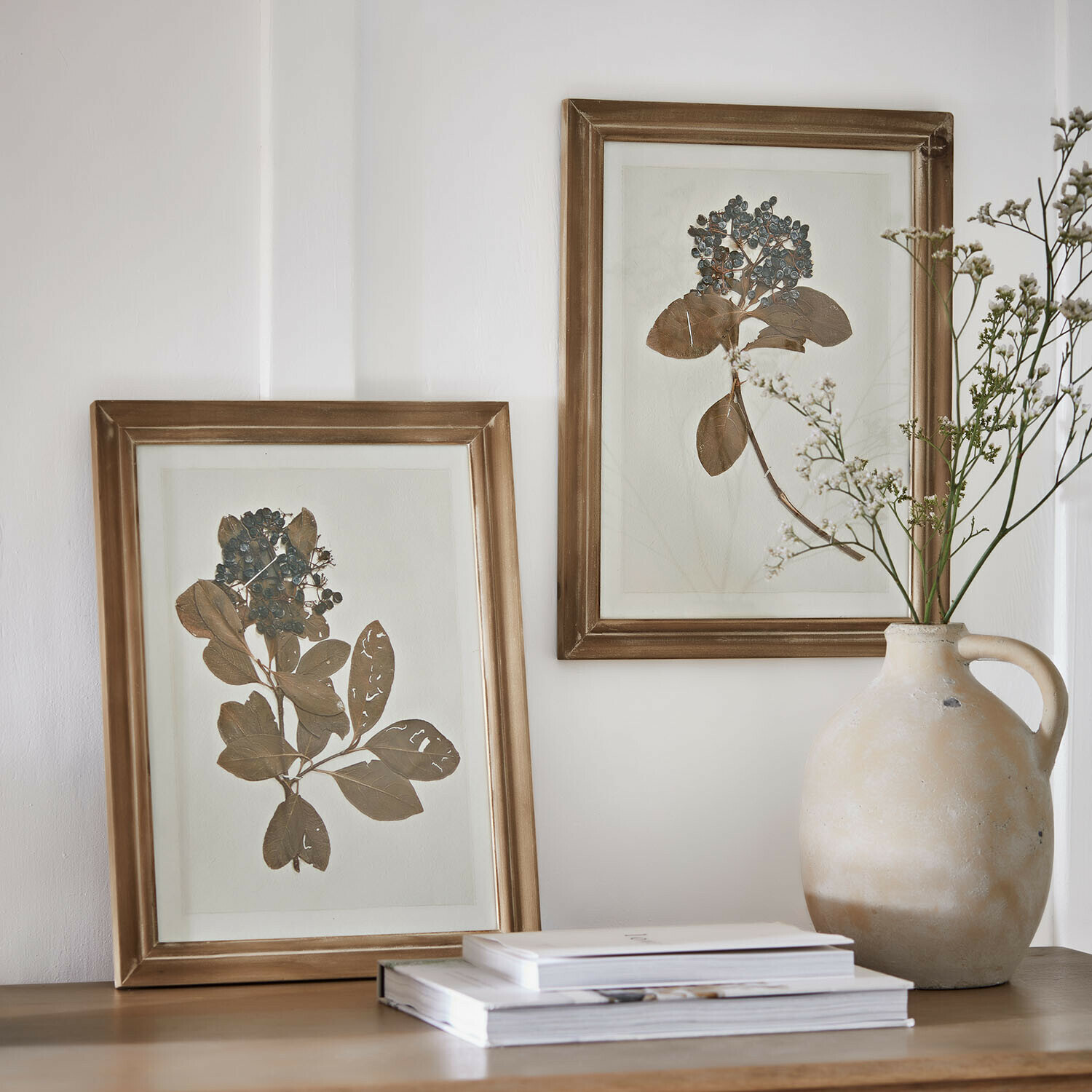 Zwei goldene Bilderrahmen mit getrockneten Blättern und Beeren auf Holztisch neben Keramikvase und Büchern