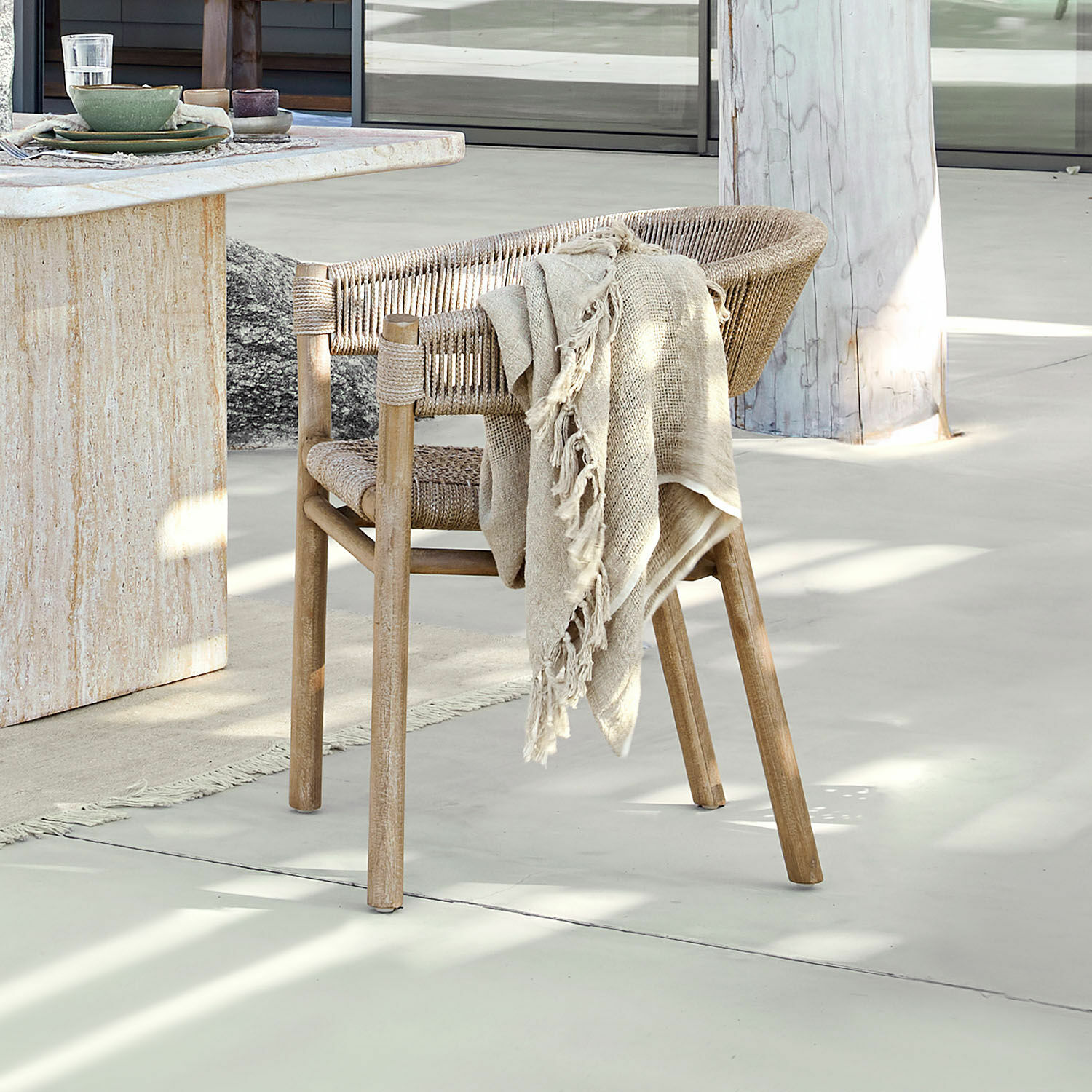 Stuhl FSC®-100% Garmontique Stuhl aus Holz und Naturseil in hellem Farbton mit beigem Plaid vor hellem Boden und Tisch.