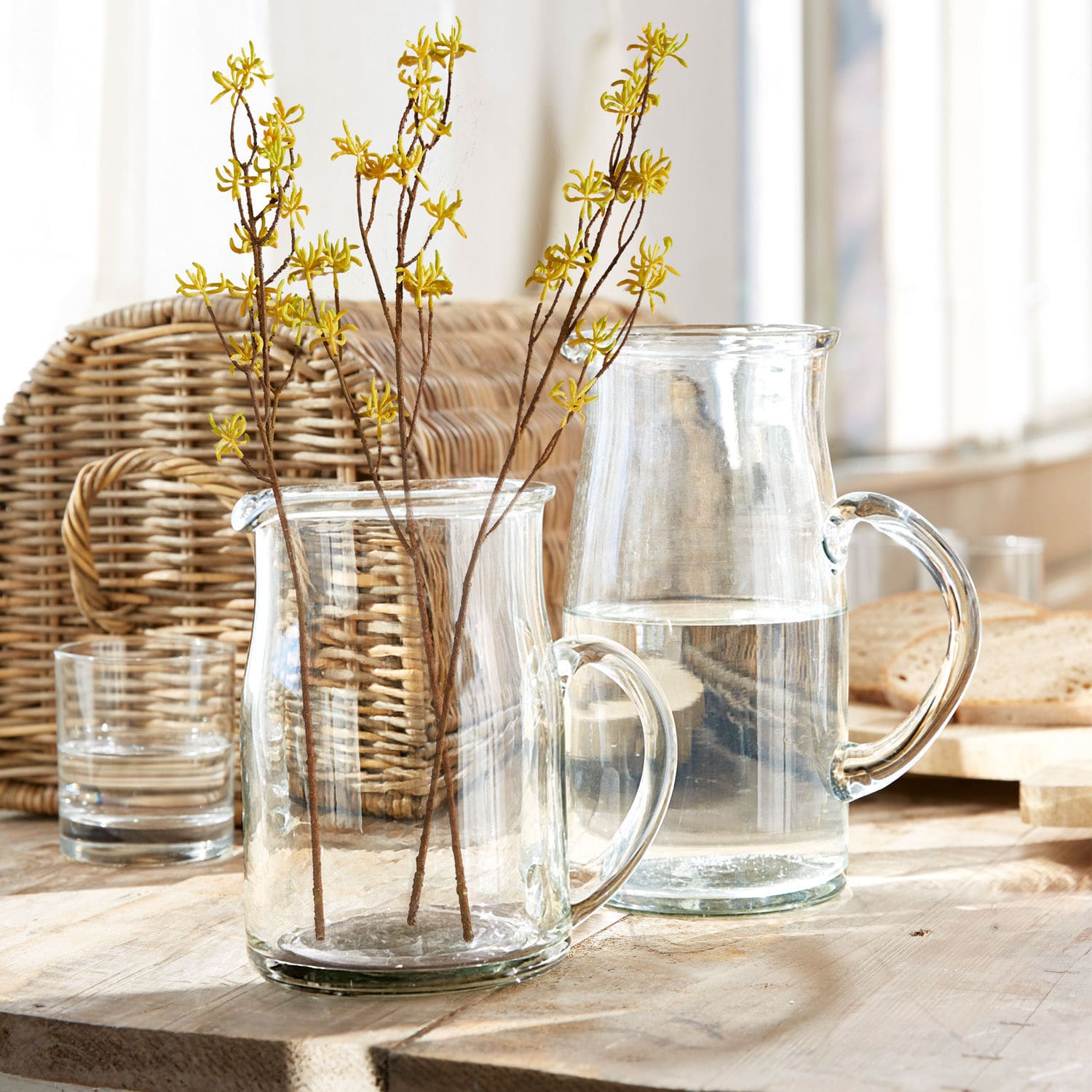 Transparenter Glaskrug und Krug mit gelben Blütenzweigen auf hellem Holztisch