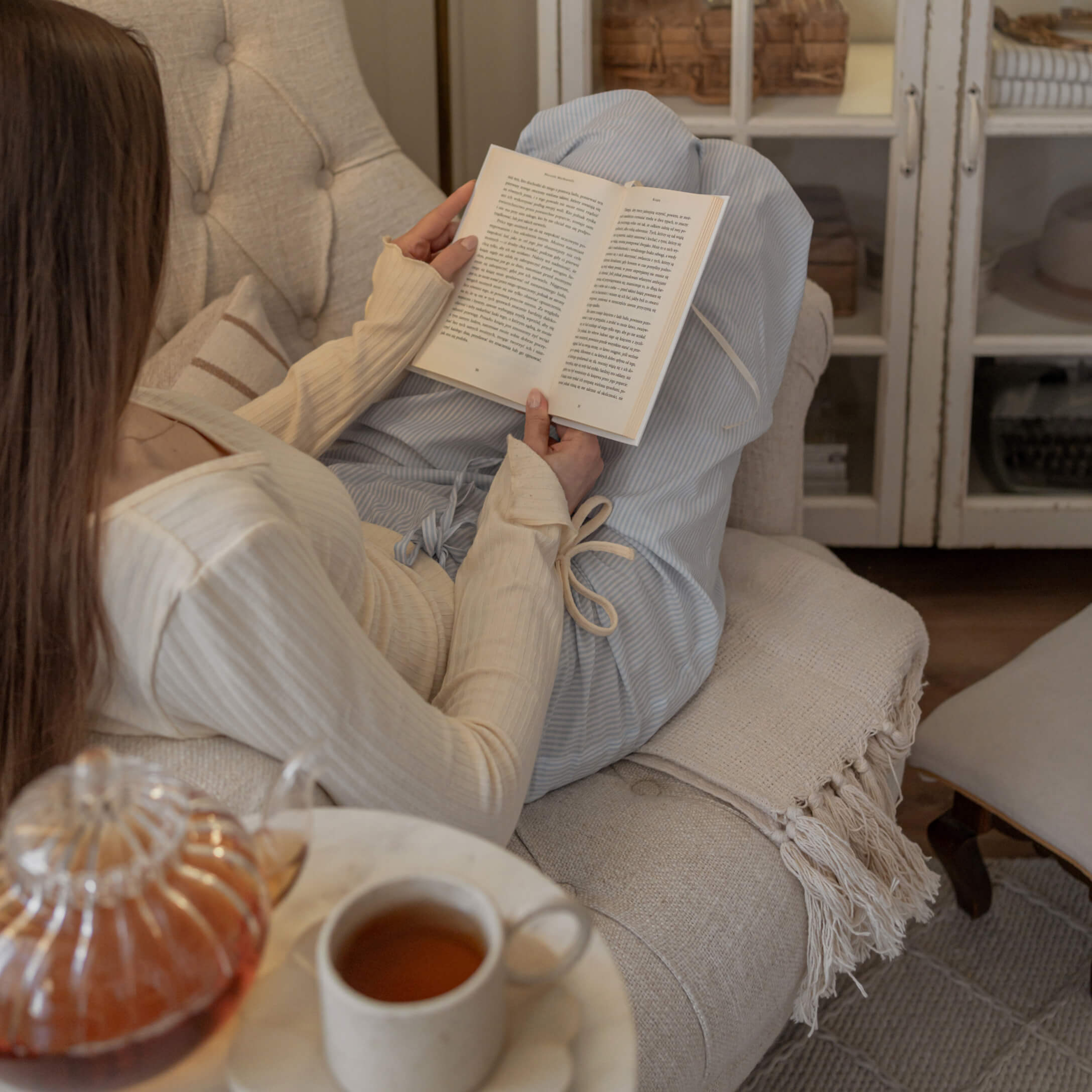 Frau in cremefarbenem Pullover und blauer Hose liest Buch auf beigem Sofa mit Tee auf dem Tisch