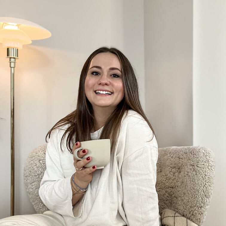 Lächelndi Frau im wiisse Pullover hält e beige Tasse, sitzt uf eme weiche Sessel i hellem Raum