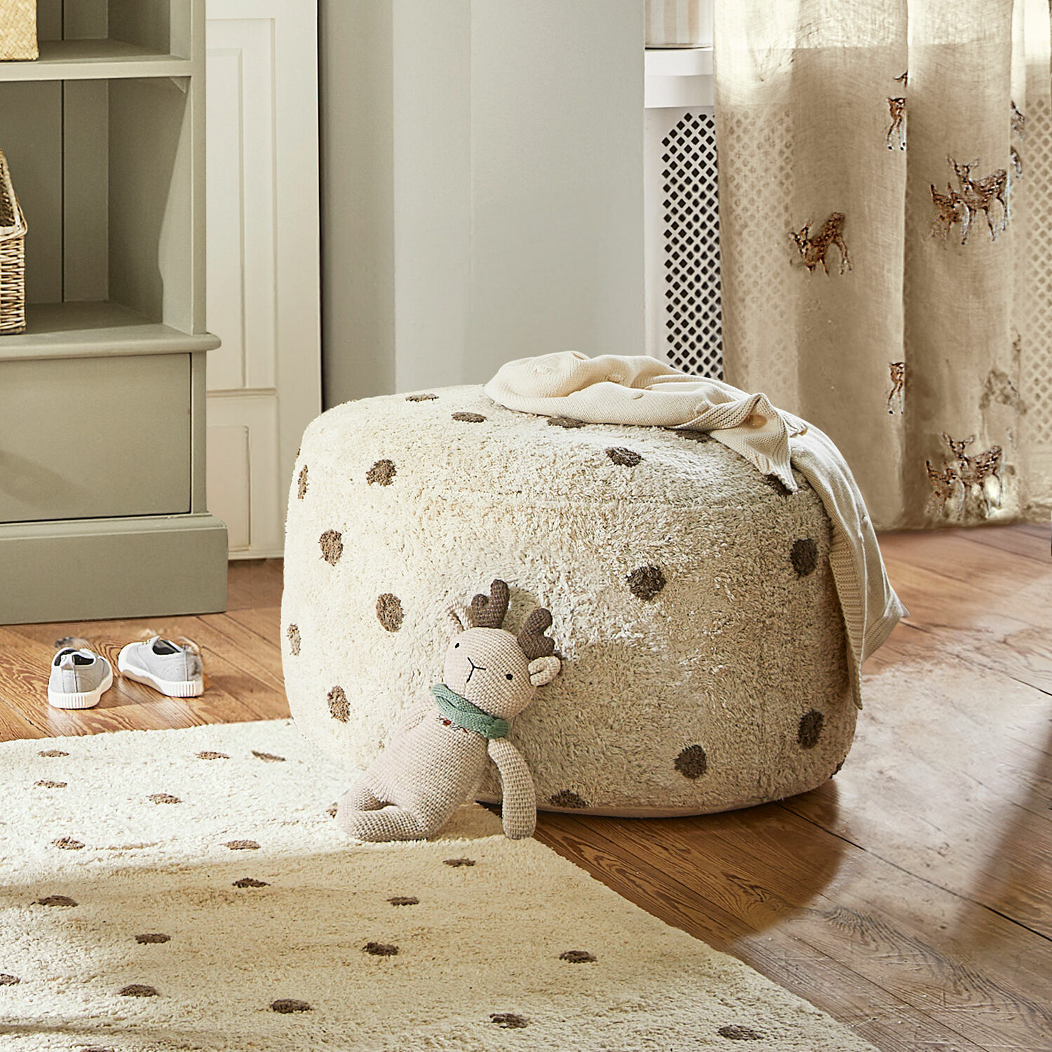 Pouf Thivalie Beiger, weicher Polsterhocker mit Punkten auf Holzboden, Kuscheltier und Teppich im Raum.