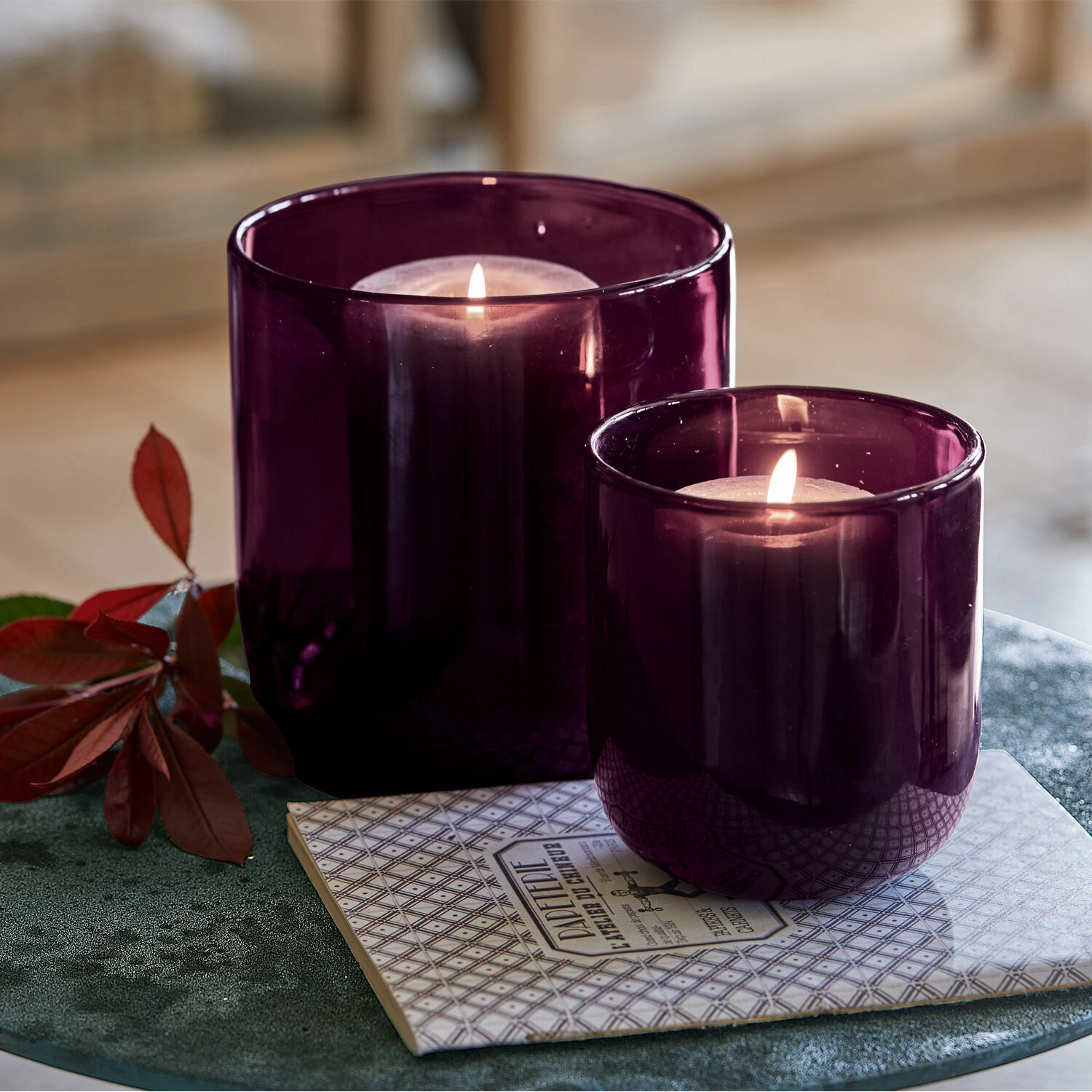 Windlicht 2er Set Theodor Zwei violette Glaswindlichter mit brennenden Kerzen auf dekorativer Unterlage auf grünem Tisch.