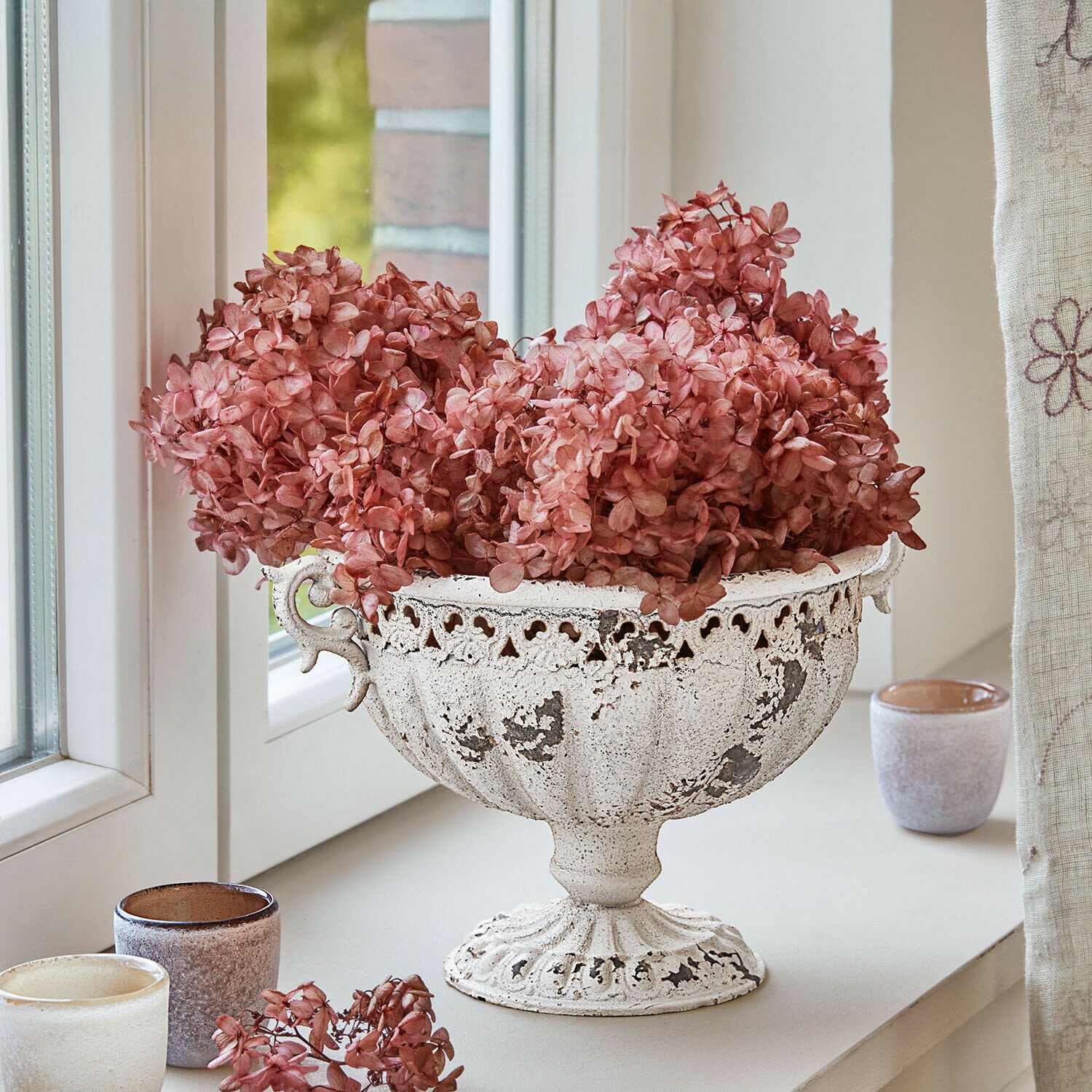 Weiße Metallschale mit antikem Finish und getrockneten rosa Blumen auf Fensterbank