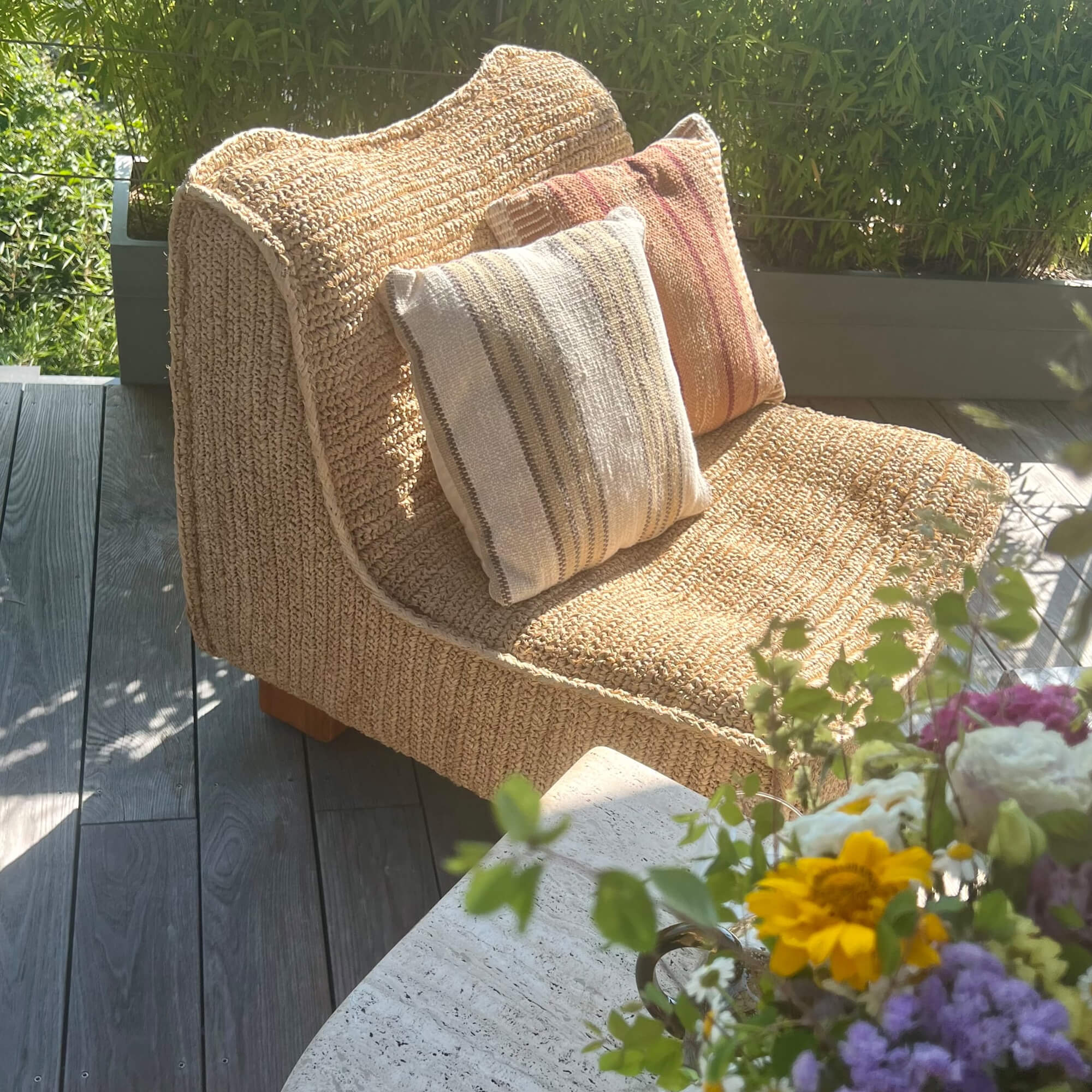 Beiger Sessel aus geflochtenem Material mit zwei dekorativen Kissen auf der Terrasse mit Blumen