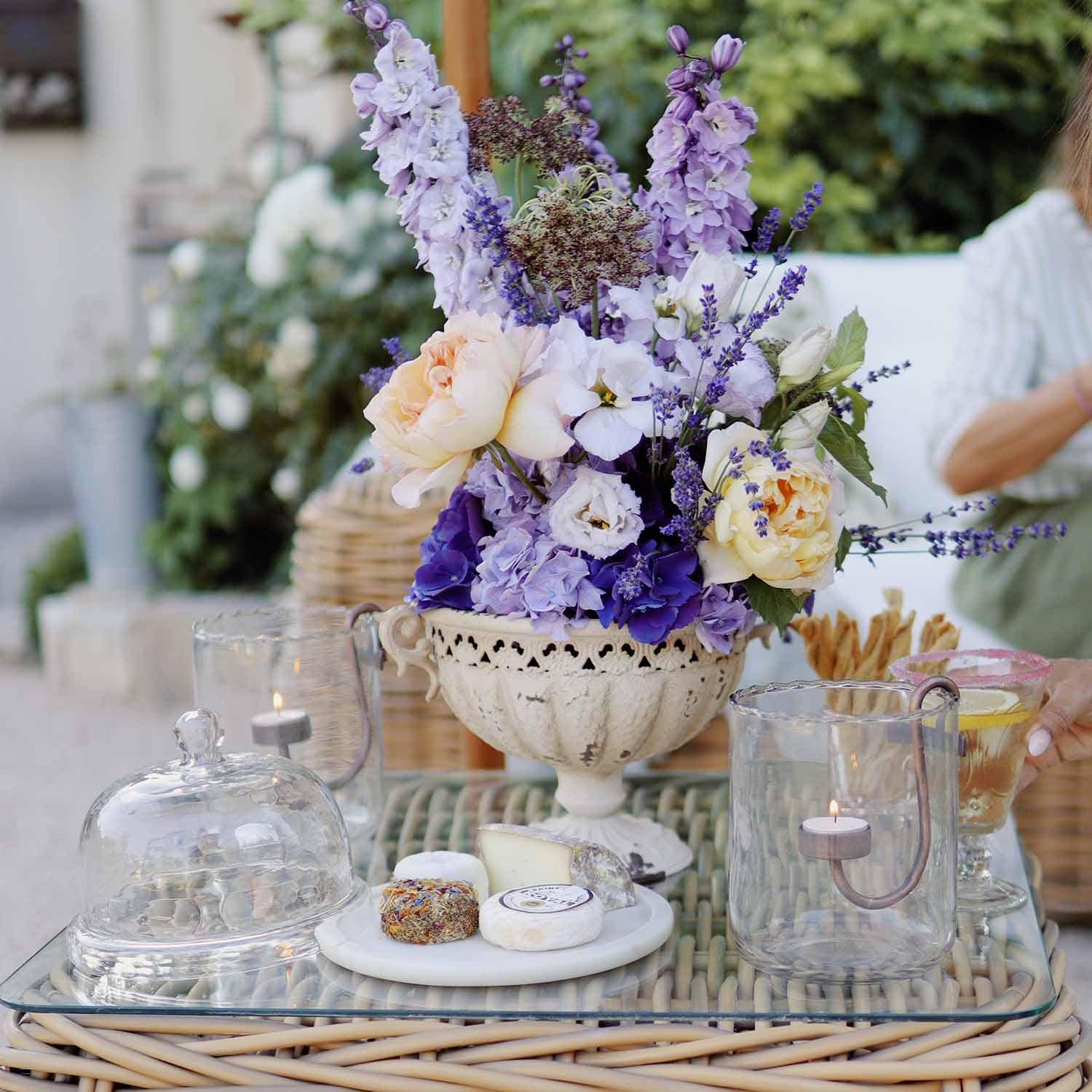 Strauß aus lila und cremefarbenen Blumen in weisser Keramikvase auf Glastisch mit Kerzen und Käseplatte