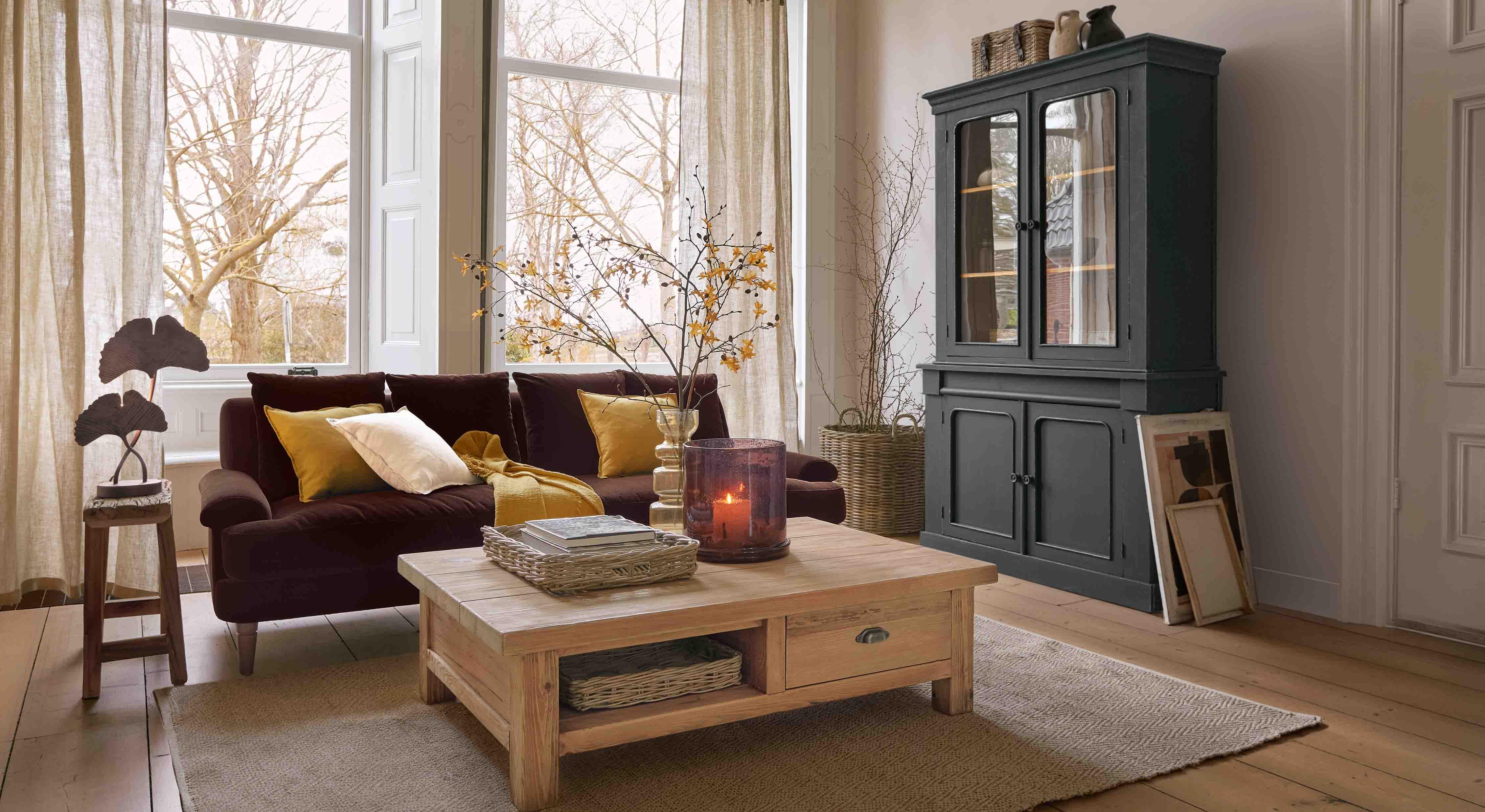 Gemütliches Wohnzimmer mit dunklem Sofa, Holztisch und schwarzem Holzvitrinenschrank vor großen Fenstern
