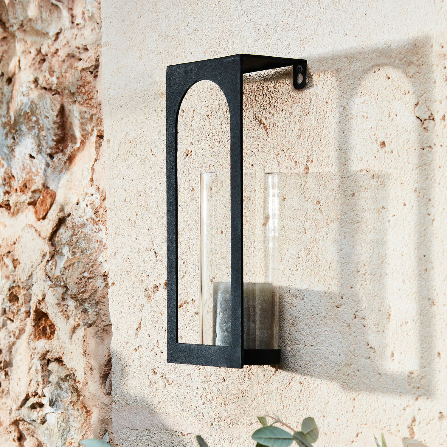 Wandkerzenhalter Maidis Schwarze Metall-Wandleuchte mit klarem Glaszylinder und Kerze an beiger Steinwand