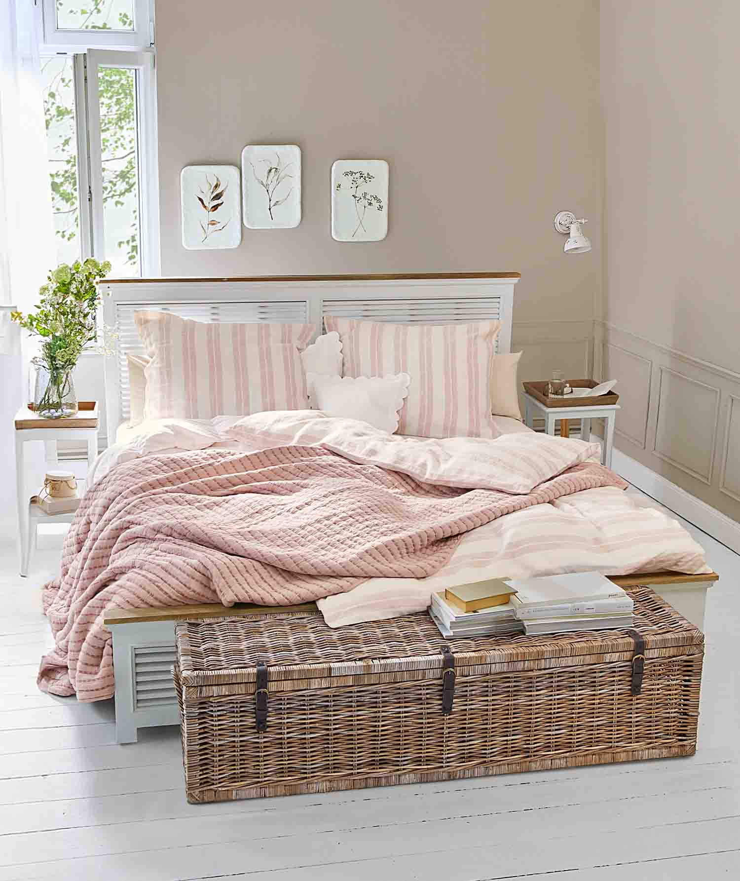 Gemütliches Schlafzimmer mit weissem Bett, rosa gesteppter Decke und Korbtruhe mit Büchern darauf