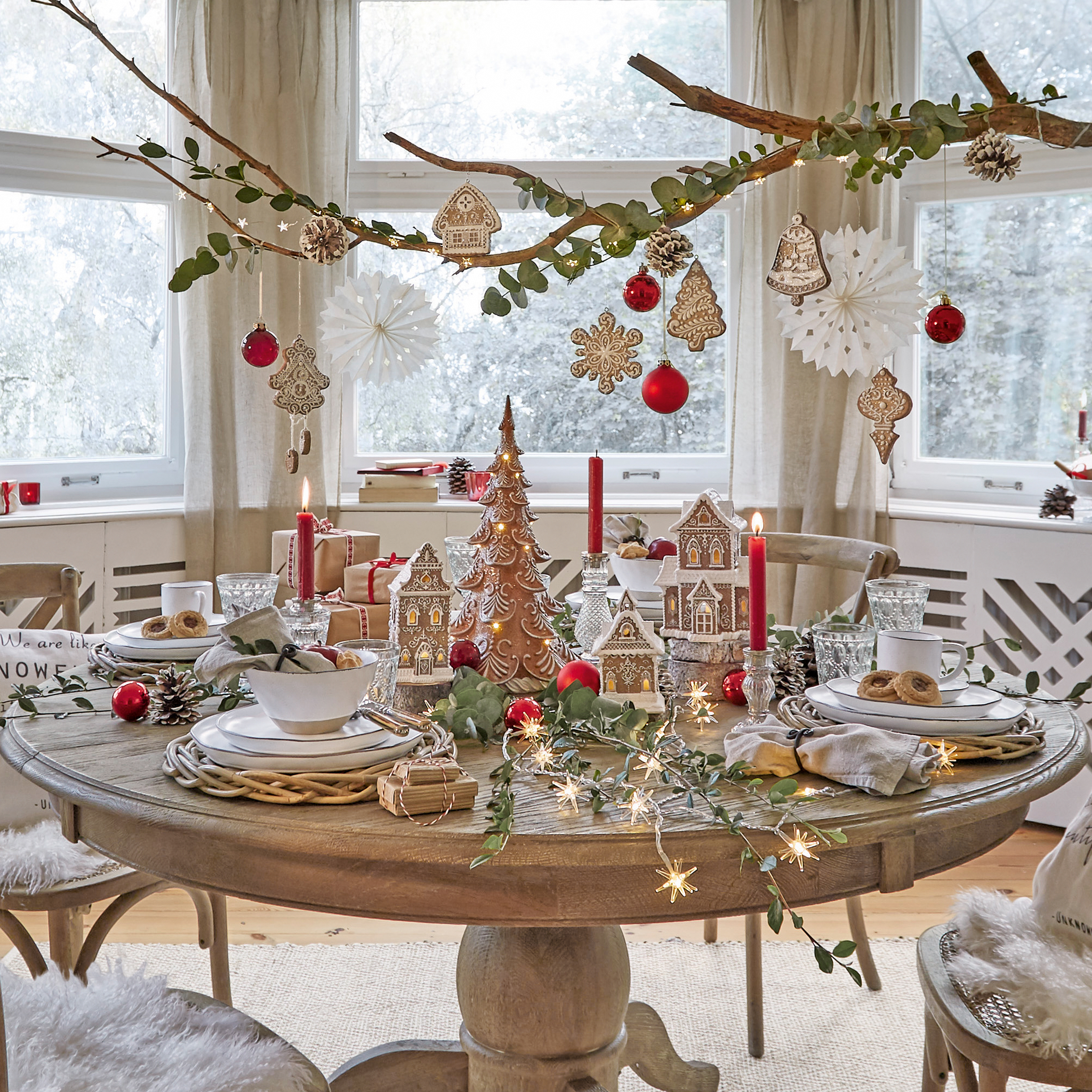 Runder Holztisch mit Weihnachtsdekoration aus Lebkuchenhäusern, roten Kugeln und brennenden Kerzen