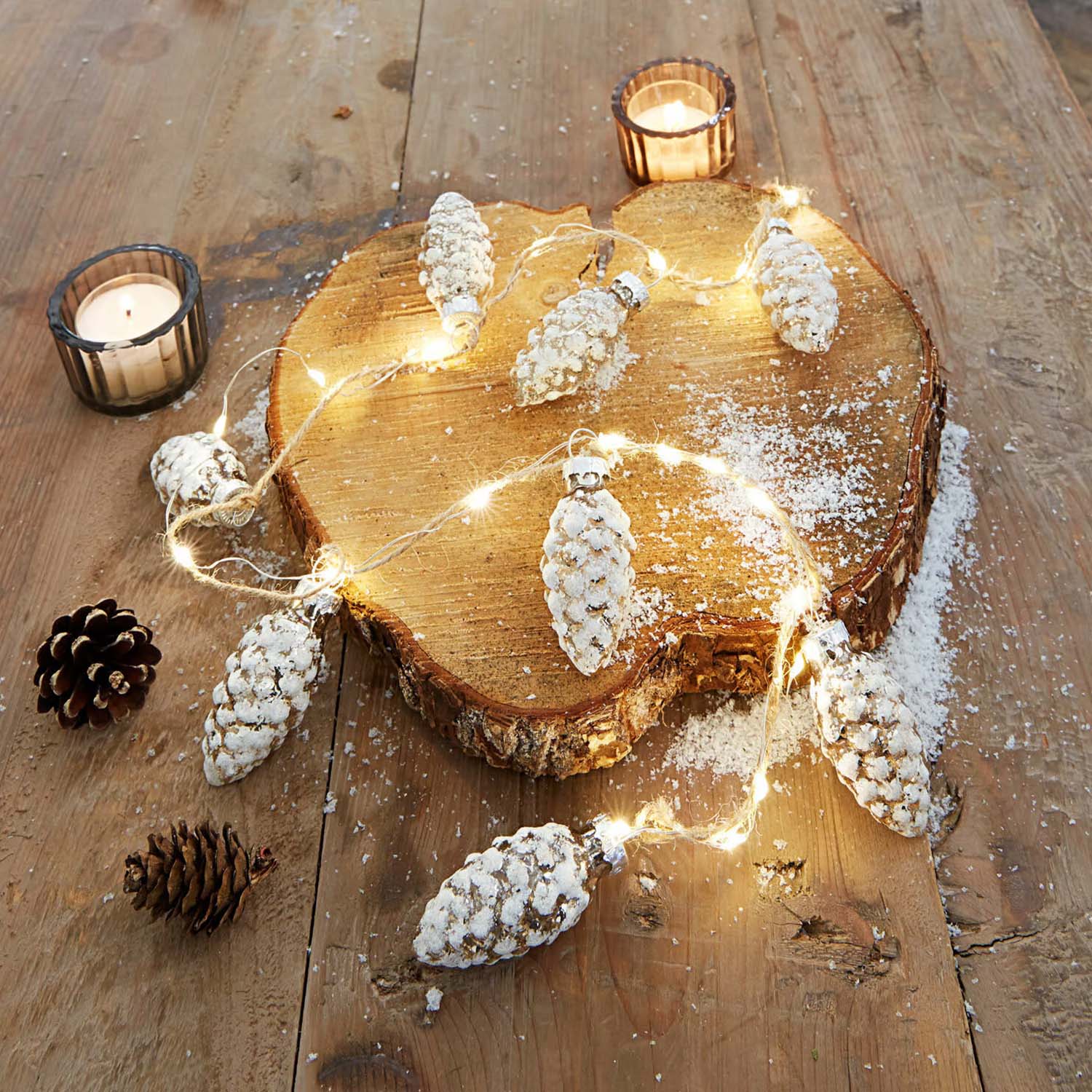 Lichterkette mit schneebedeckten Zapfen auf Holzscheibe mit zwei Kerzen auf rustikalem Holztisch