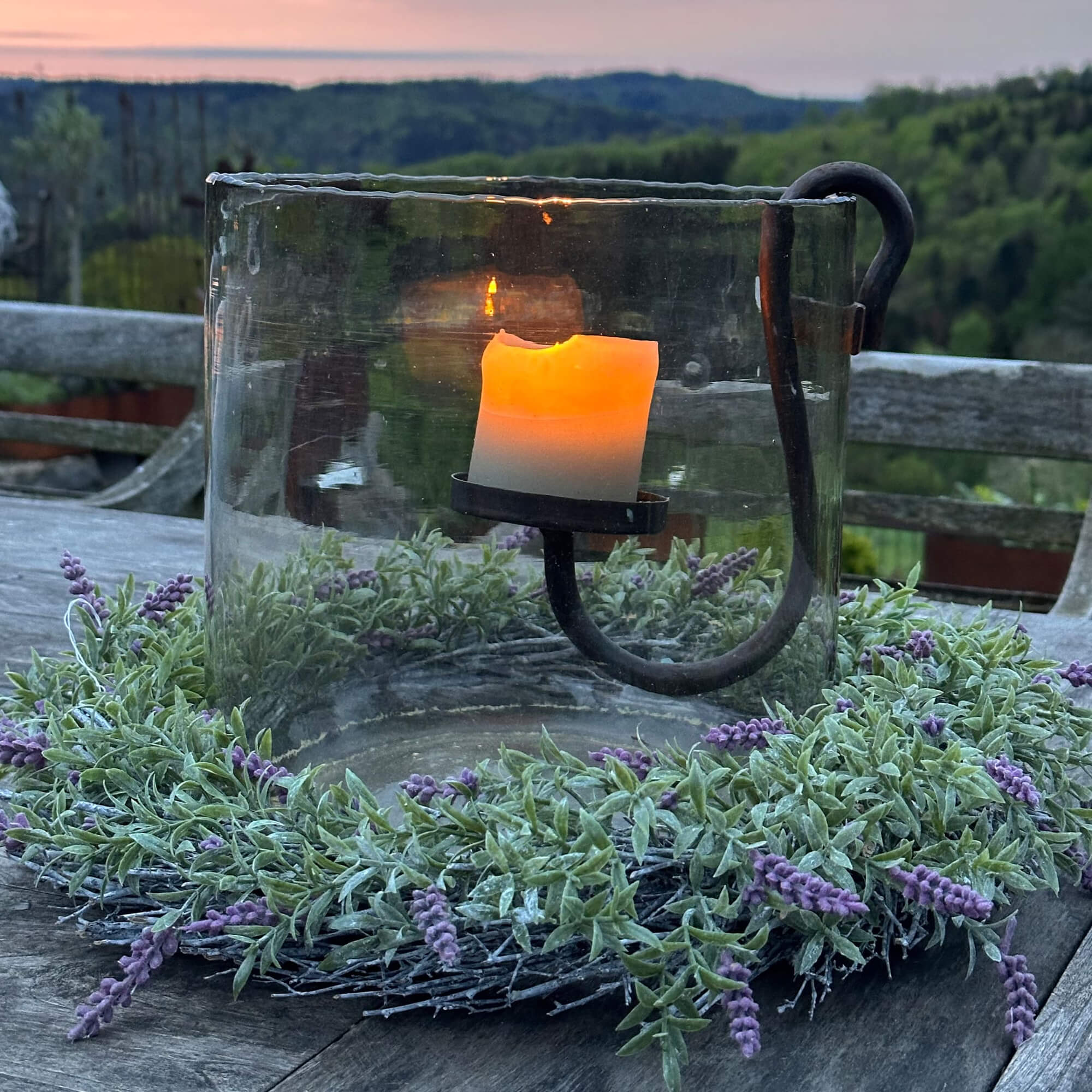 Glaswindlicht mit Metallhalterung und grünem Kranz mit violetten Blüten auf Holztisch