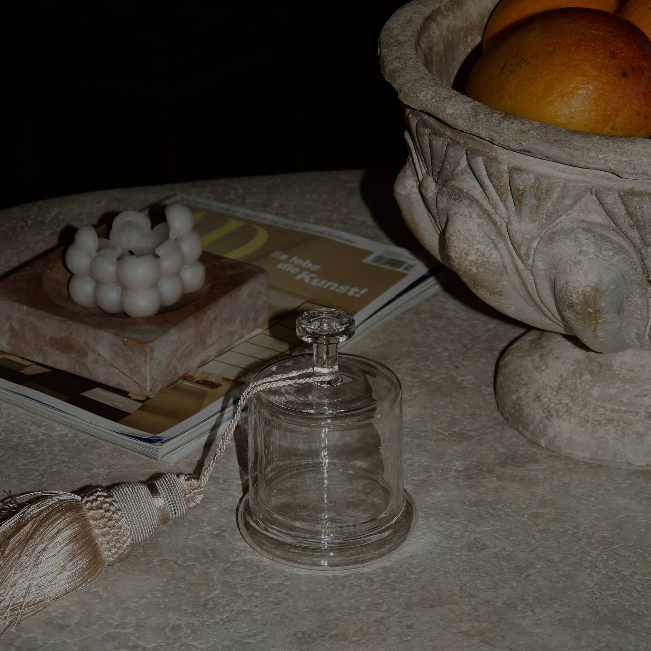 Transparente Glasglocke mit dekorativem Quastenanhänger auf Marmorplatte mit Kerze und Steinschale