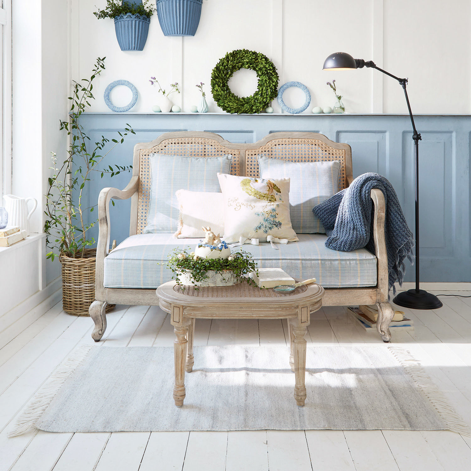 Sofa Ainsley Holz-Rattan-Sofa mit blauer Polsterung und Kissen im hellen Wohnzimmer mit Pflanzen und Stehleuchte