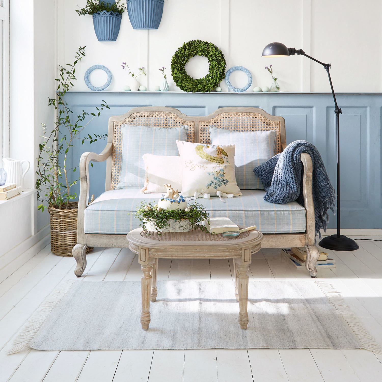 Sofa Ainsley Holz-Rattan-Sofa mit blauer Polsterung und Kissen im hellen Wohnzimmer mit Pflanzen und Stehleuchte
