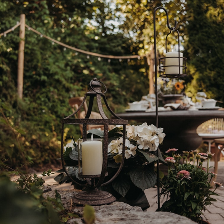 Metalllaterne mit weisser Kerze auf Steinmauer im Garten mit Blumen und Tisch im Hintergrund