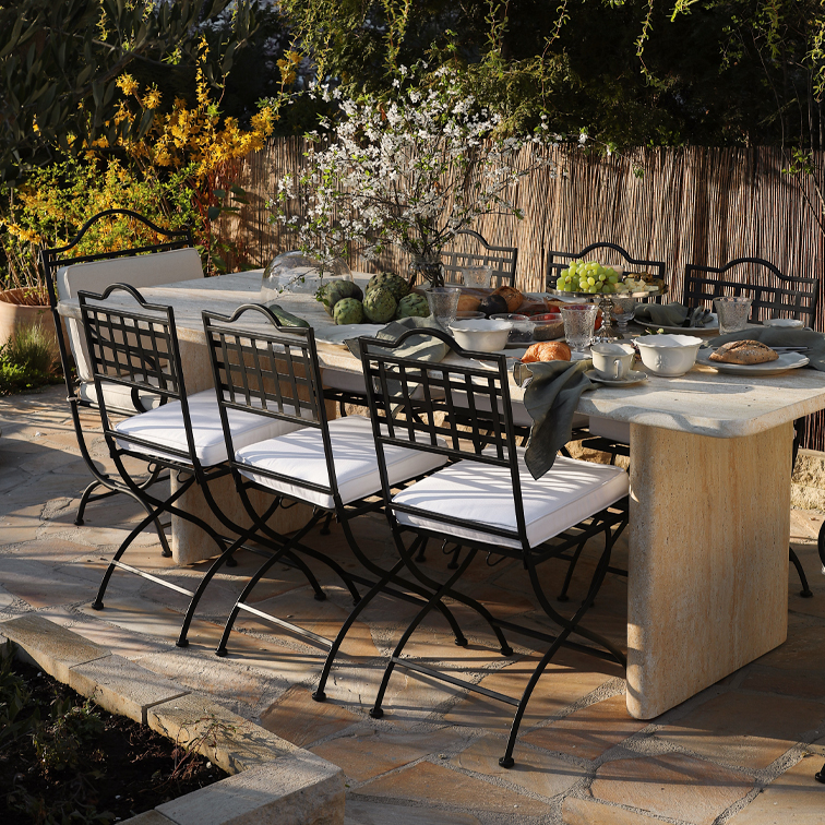 Gartentisch aus Stein mit schwarzen Metallstühlen und weissen Sitzkissen, gedeckt für eine Mahlzeit im Freien