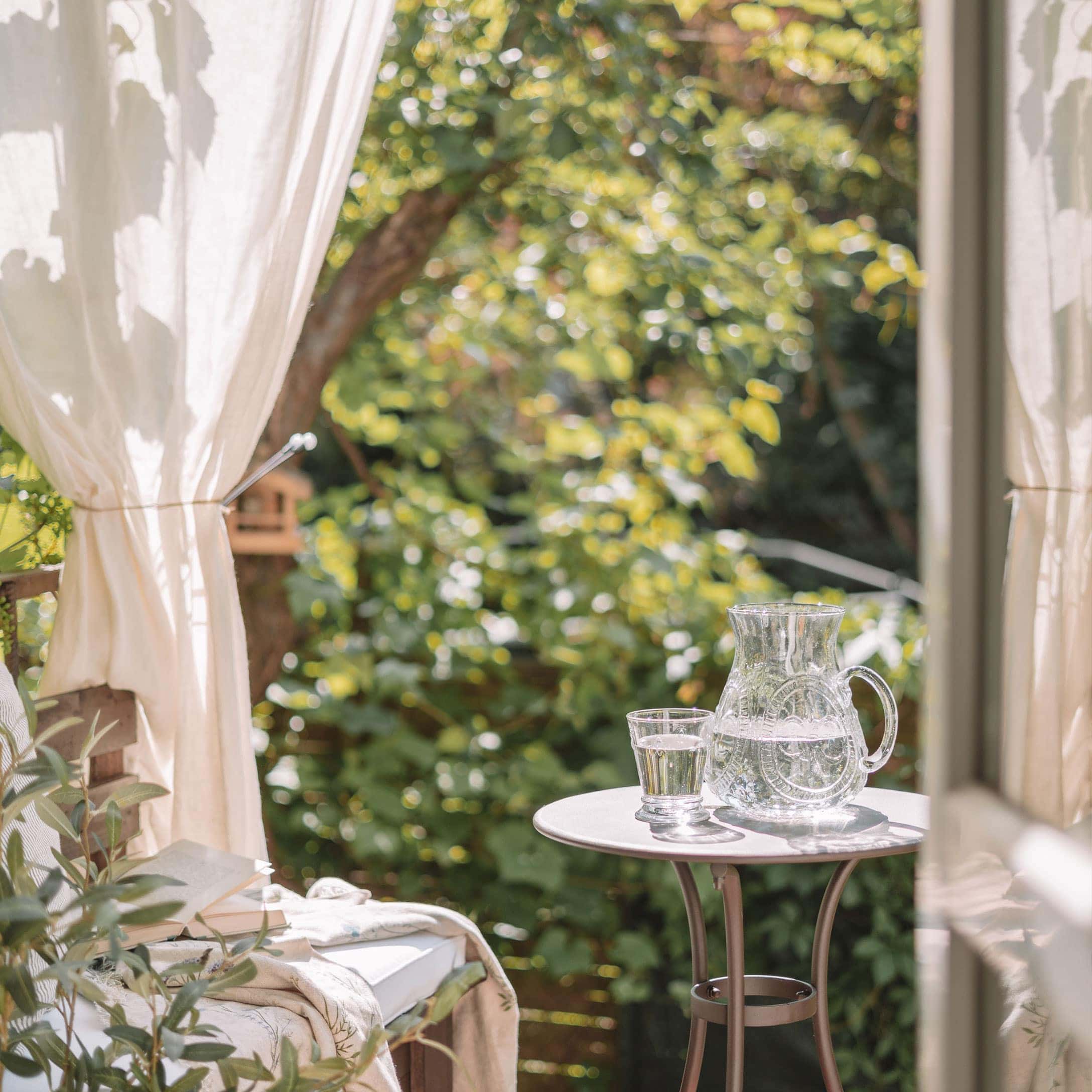 Glaskrug und Glas mit Wasser auf weissem Tisch auf Terrasse mit grünem Pflanzenhintergrund