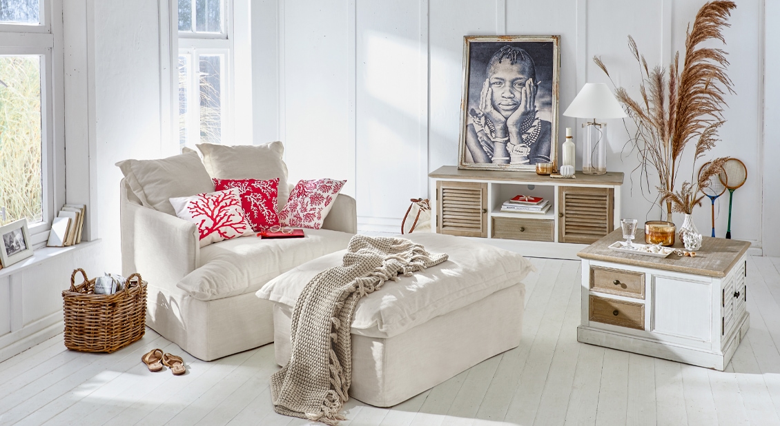 Helles Wohnzimmer mit beigem Sofa, roten Kissen und weissen Holzmöbeln im Strandhausstil