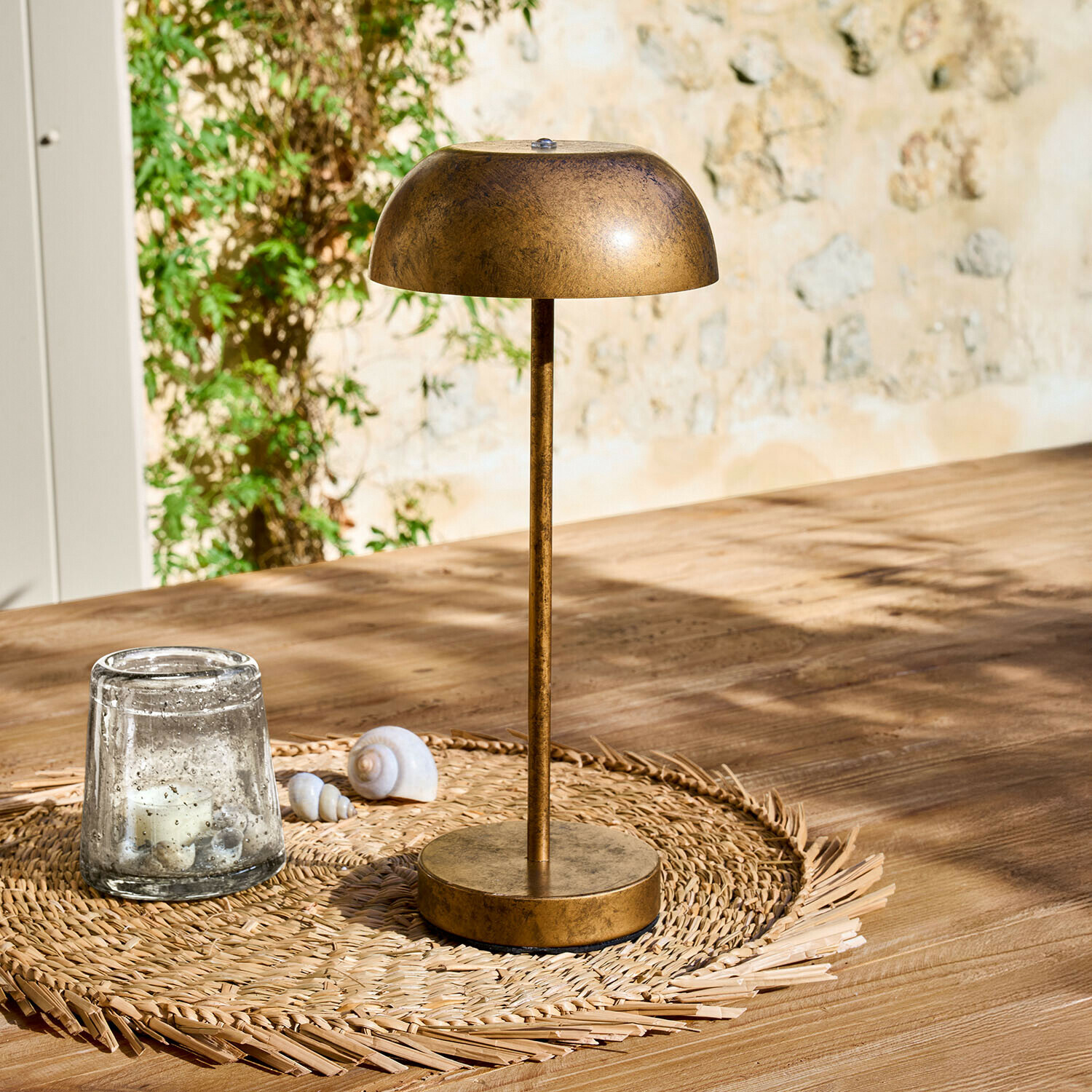 Tischlampe aus Metall in antikgold auf geflochtenem Untersetzer auf Holztisch im Sonnenlicht