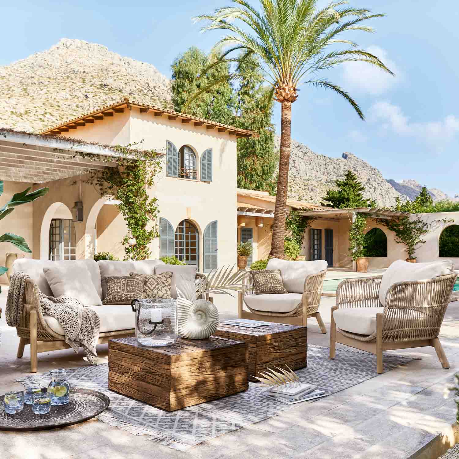 Outdoor-Möbelset aus hellem Rattan mit beigen Polstern auf Terrasse mit Palme und Bergsicht