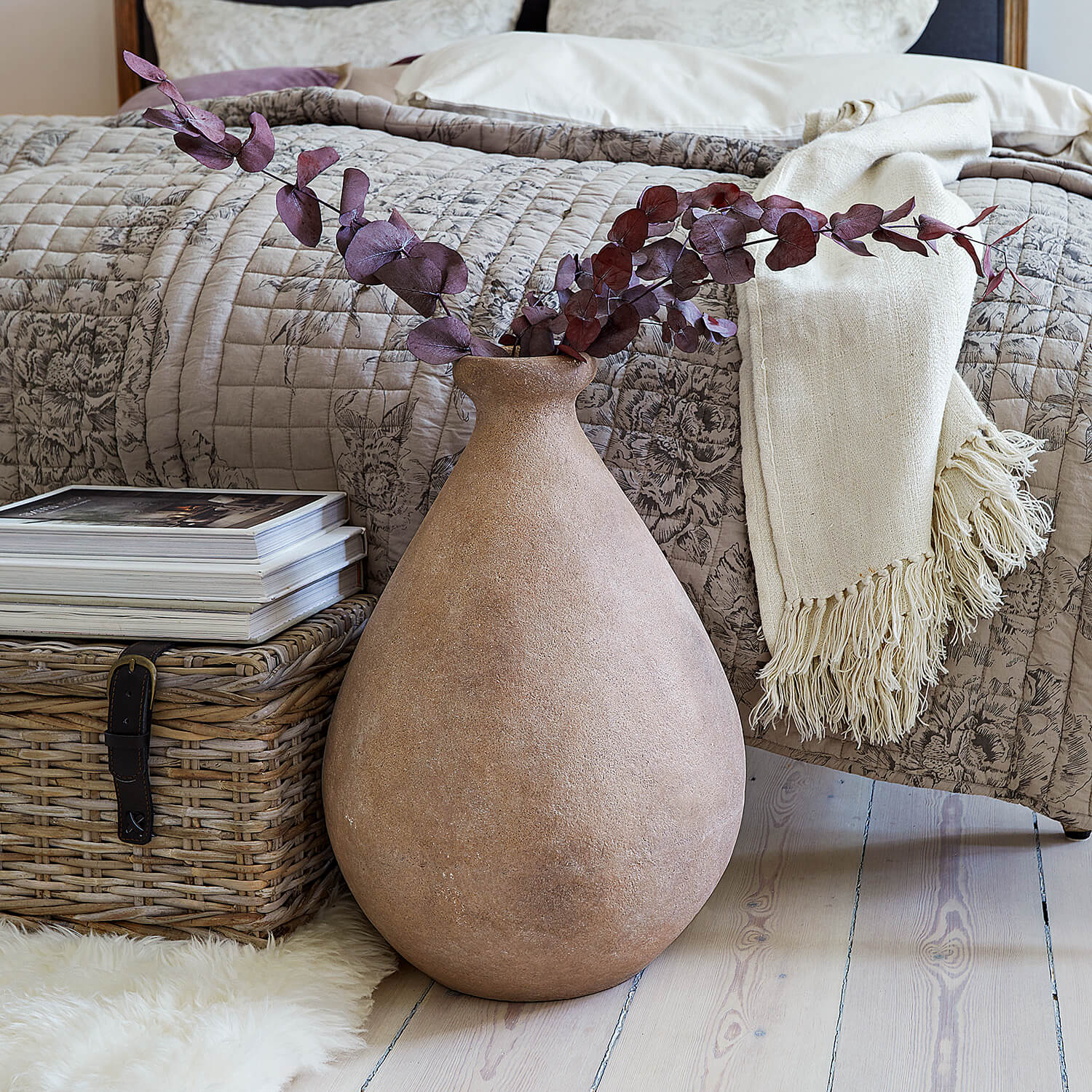 Grosse beige Keramikvase mit getrockneten Zweigen vor Bett und Weidenkorb